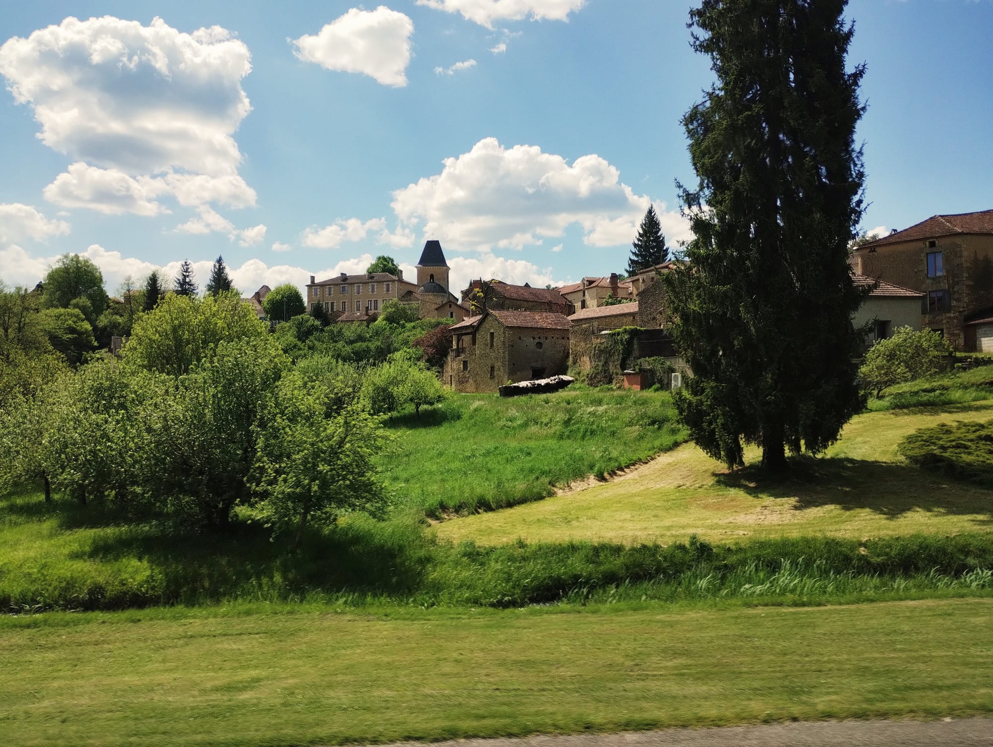 A view of a village in the south-west of France. Upon a hill there is a church tower and a large building. There are stone houses with Roman tiles beneath it and lush grass and trees.