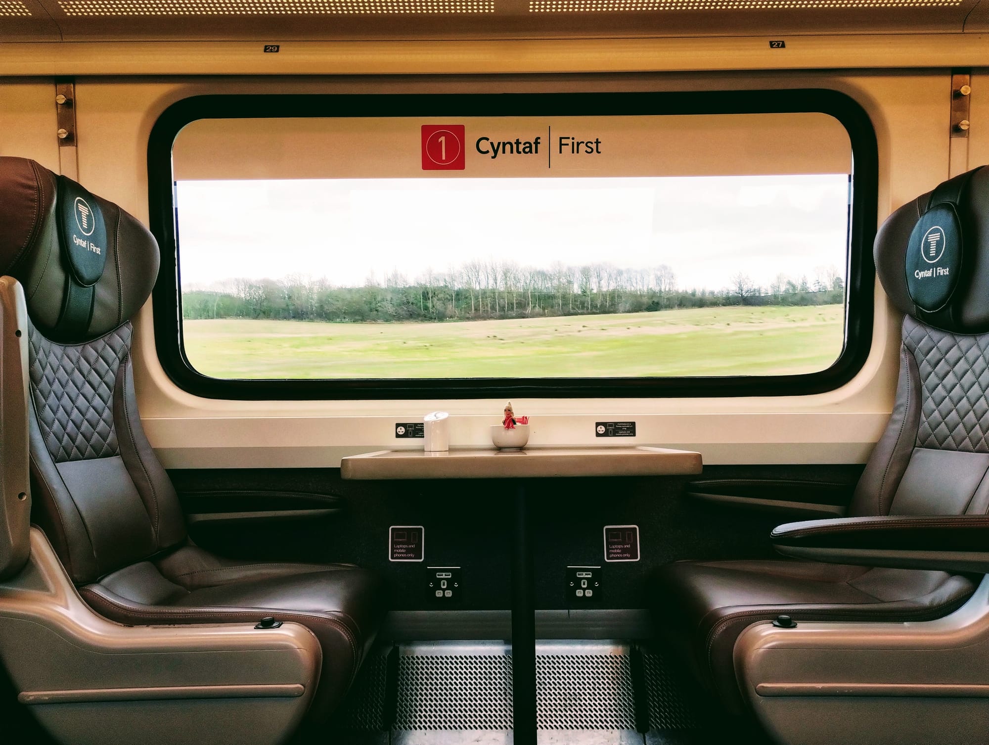 Interior view of a first-class train carriage with two leather seats facing each other across a small table, framed by a large window with a view of a green field and trees under a cloudy sky. The seats have diamond-stitched detailing and headrests with "Cyntaf | First" branding. A small table between the seats holds a small, white bowl containing a finger puppet.
