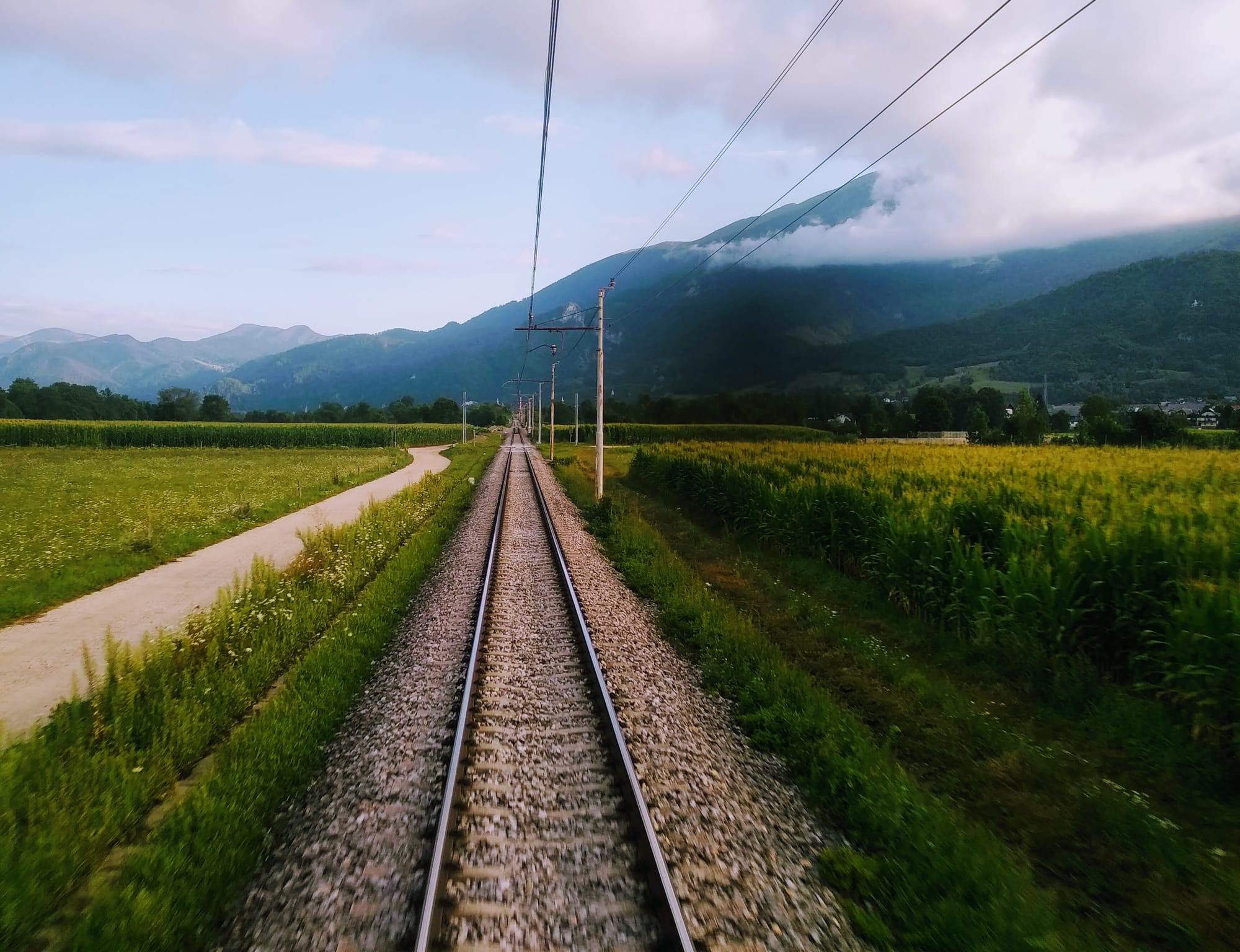 Train tracks stretching into the distance, flanked by green fields and a mountain range in the background, under a cloudy sky. Power lines run parallel to the tracks.