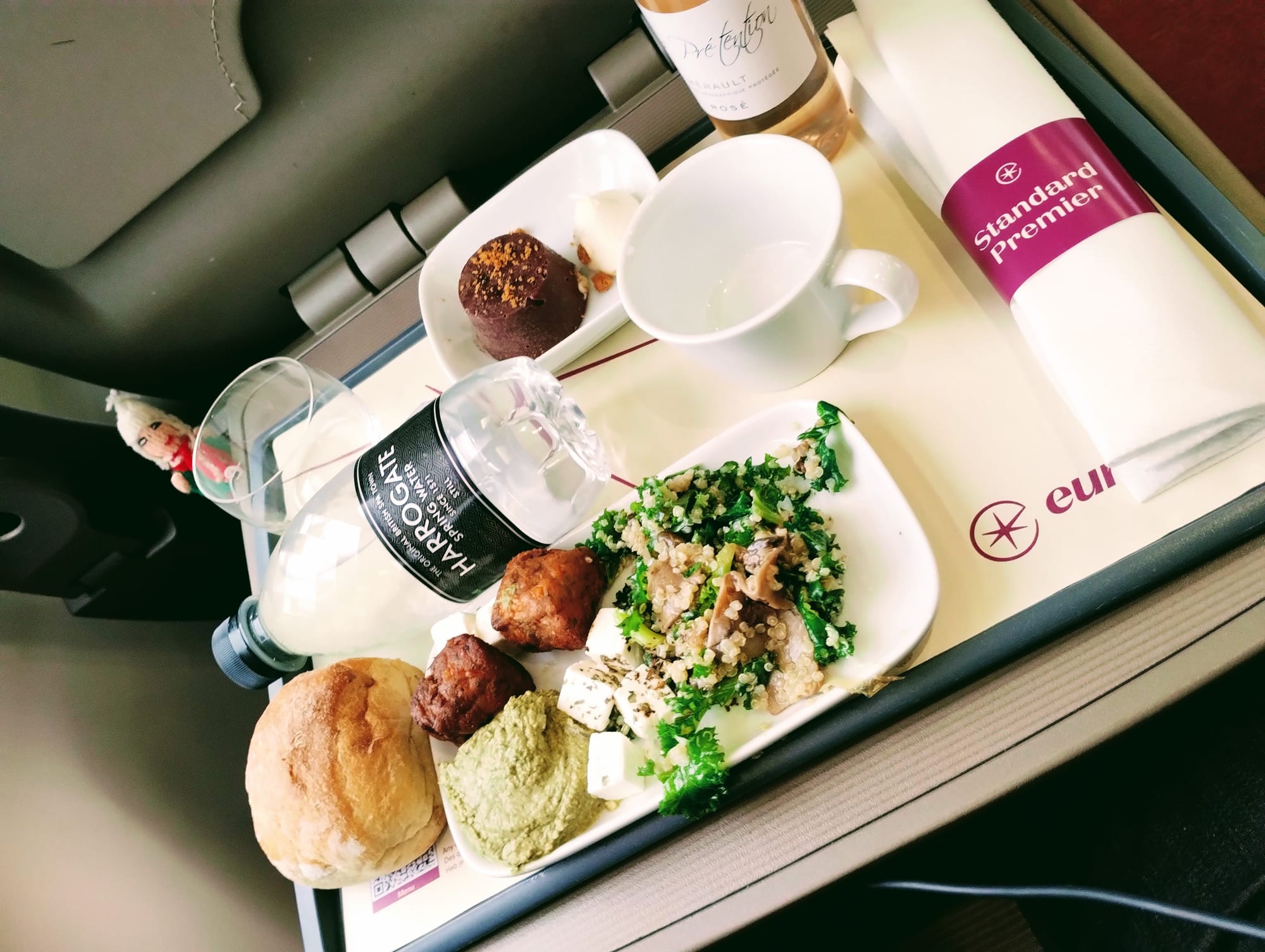 A close-up of meal tray featuring a white plate with a kale salad, falafel, hummus, and feta cheese, a bread roll, a glass of water, a bottle of rose wine, a chocolate dessert with cream, a white cup, and a "Standard Premier" napkin.