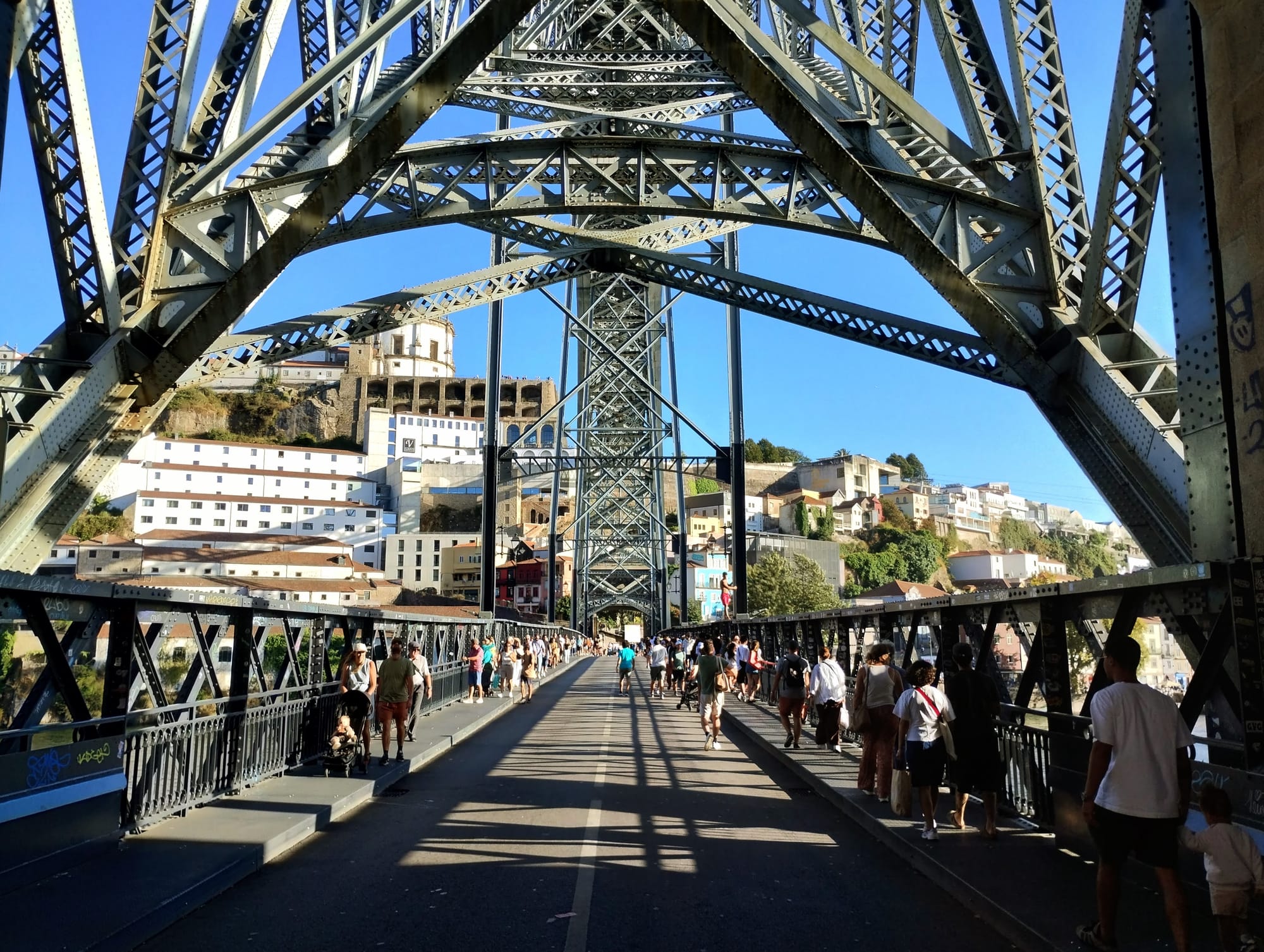 A pedestrian walkway filled with people crossing under a complex web of metal girders against bright blue sky, with buildings in the background.