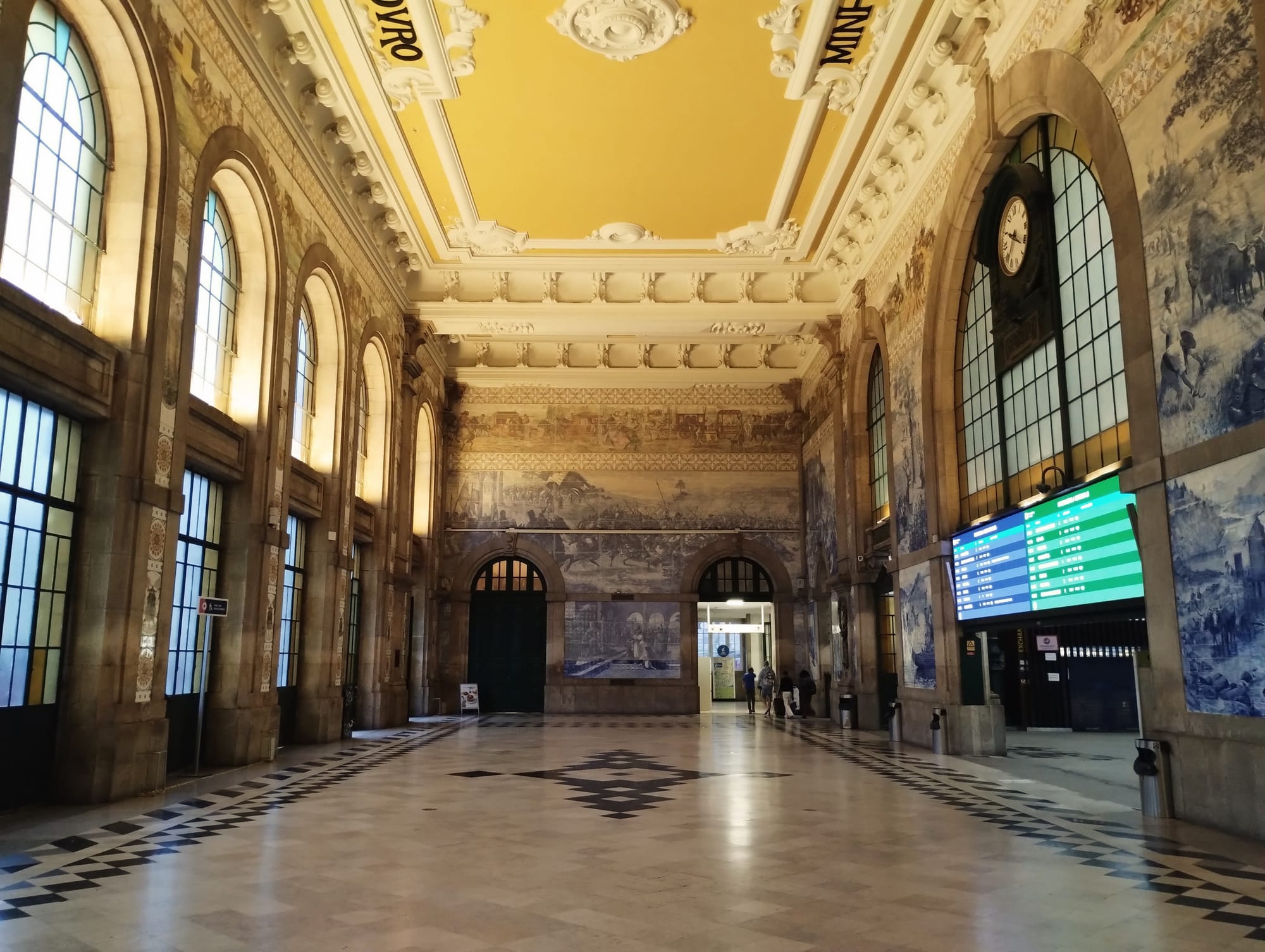 Interior of Porto Sāo Bento, featuring high arched windows, intricate ceiling moulding, and painted murals on the walls. A tiled floor in a geometric pattern. A digital display board is visible.