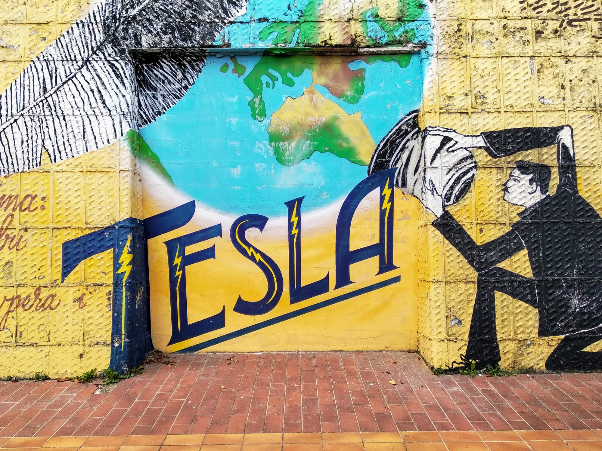 A vibrant mural on a textured yellow wall depicts Nikola Tesla next to the word "TESLA" in large blue lettering on a yellow background. On the right, Tesla holds the fitting of a light bulb formed from a depiction of the earth.