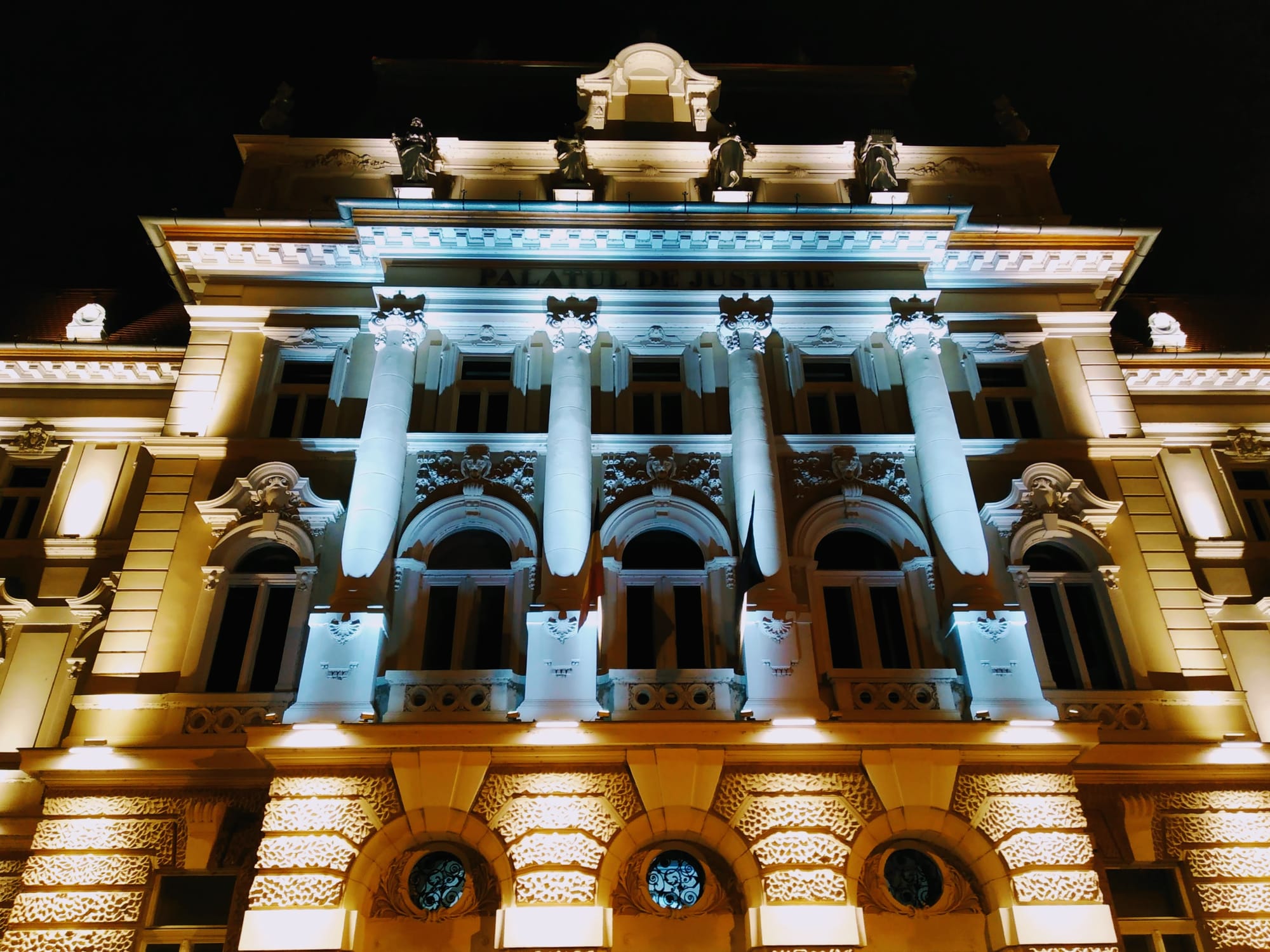 Nighttime photograph of an ornate building with architectural details illuminated by both warm and cool-toned lights. The building's facade is symmetrical, with multiple columns supporting the upper levels. A dark sky serves as the backdrop.