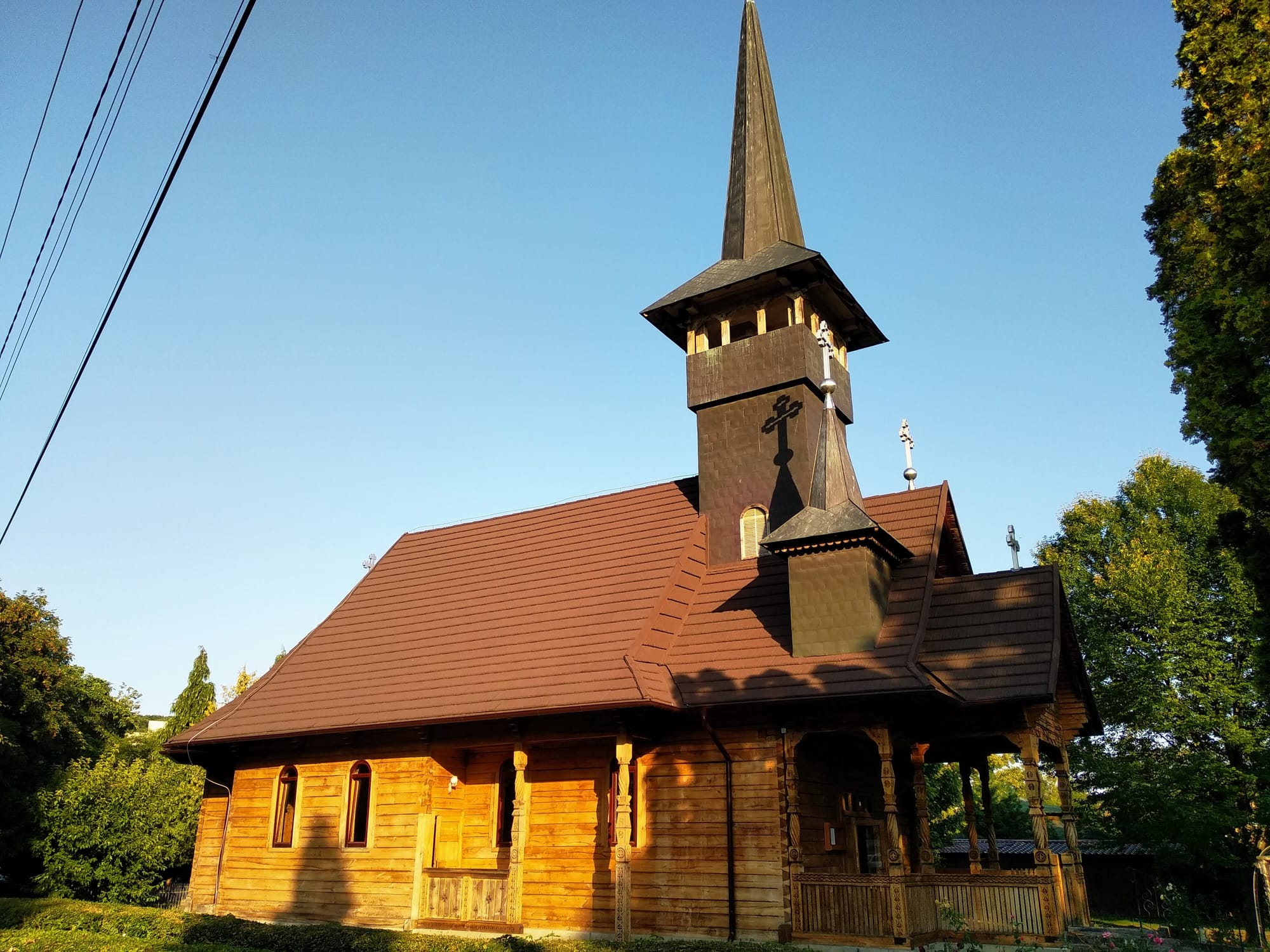 A wooden church with a tall steeple and a cross on top, brown shingled roof, and a porch with decorative pillars, against a clear blue sky.