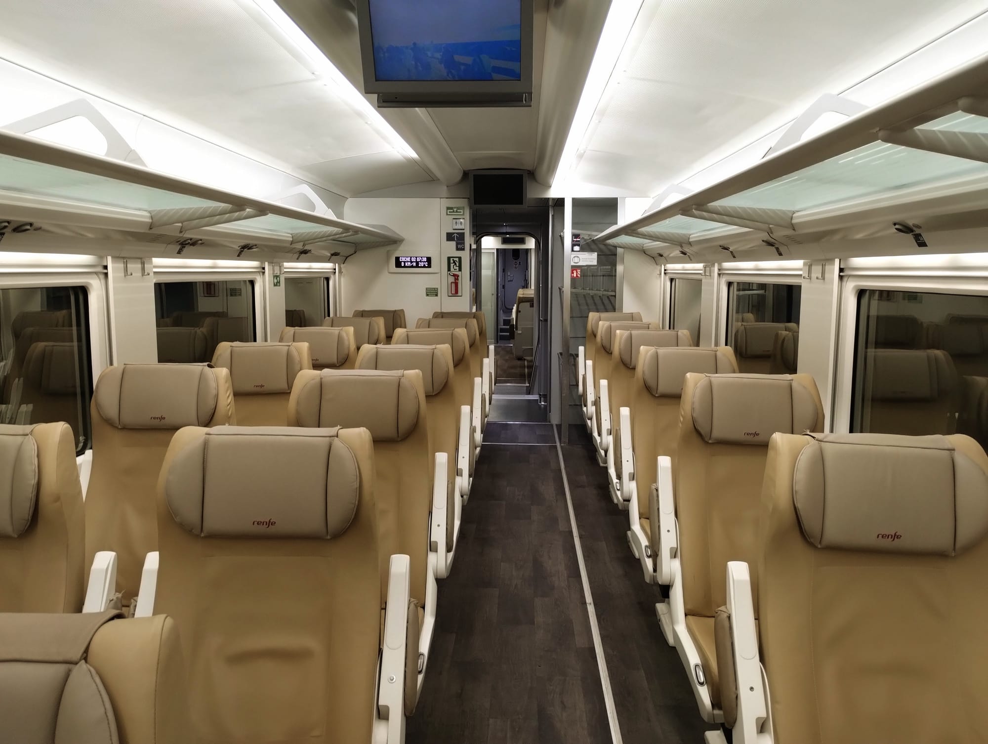 Interior view of an empty passenger train featuring rows of tan leather seats with headrests branded with "renfe". The car has overhead luggage racks, windows, and a central aisle with a door at the end.