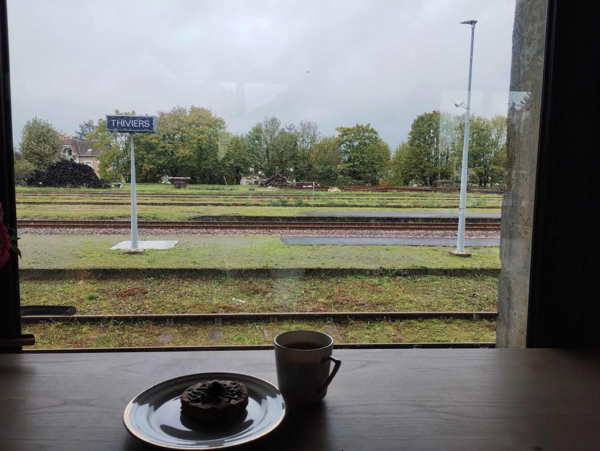 A coffee mug and a small pastry on a plate sit on a wooden table in front of a window, framing a view of the Thiviers train station under an overcast sky. The station features railway tracks, grassy areas, and a sign indicating "Thiviers."