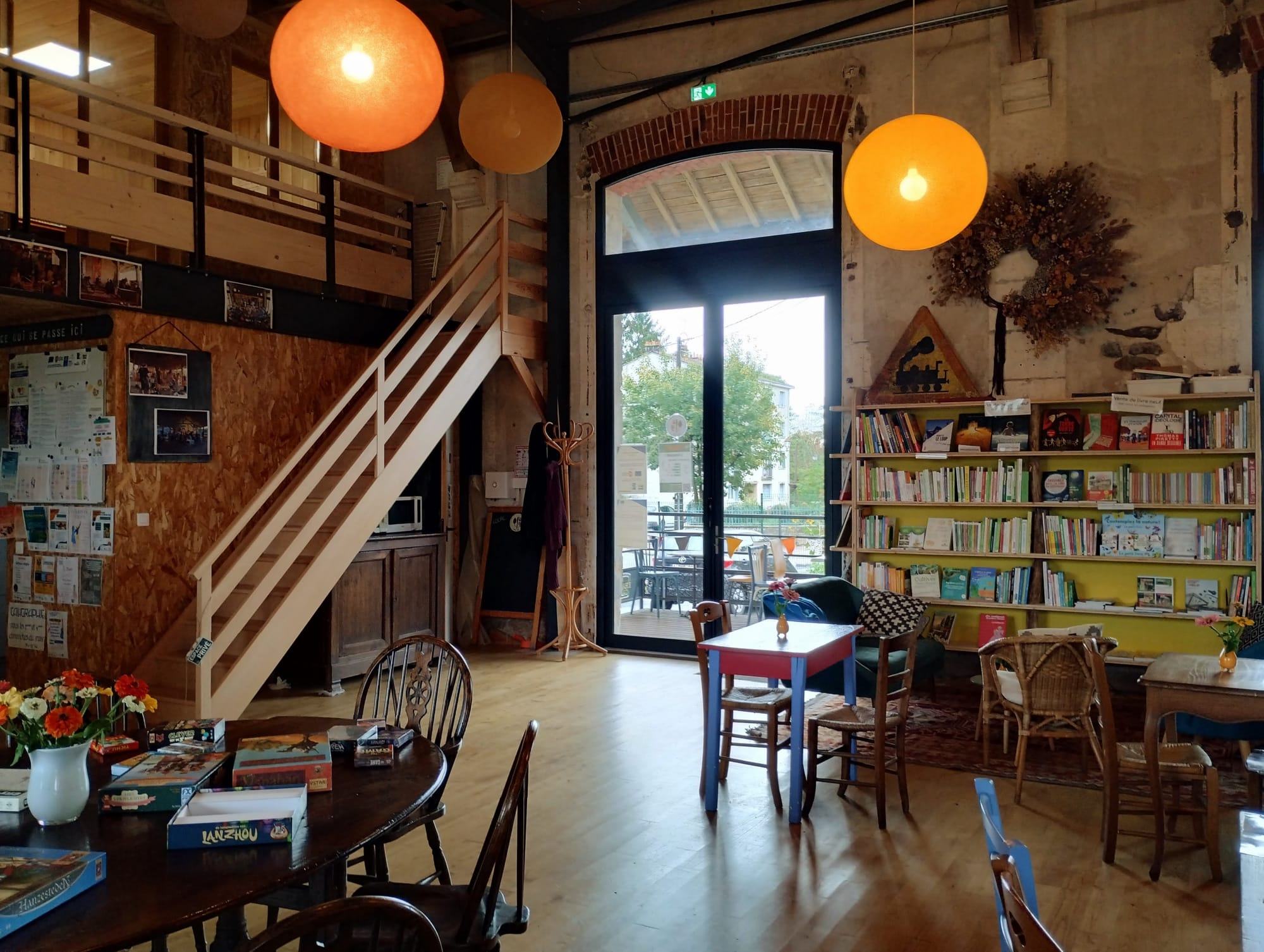 Interior view of a cafe, featuring wooden staircases leading to a loft, warm-toned globe pendant lights, bookshelves with various books, tables with board games, and a large glass door revealing an outdoor patio with additional seating.
