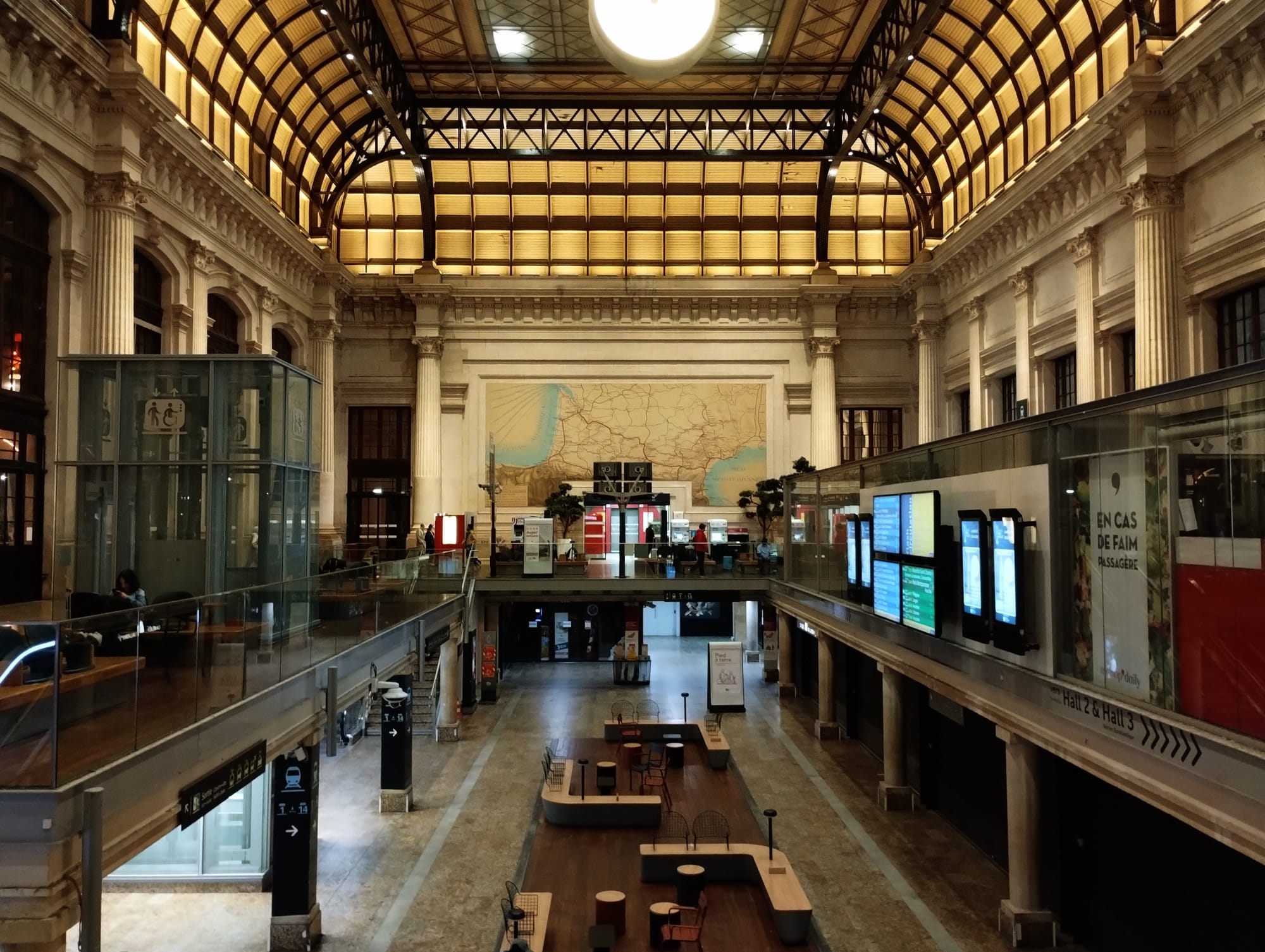 Interior view of Bordeaux Saint-Jean train station, with a high, arched ceiling with decorative lighting. The station features multiple levels, with a main floor, a mezzanine level with glass railings, and a lower level with seating areas. Large ornate columns, a map on the wall, and information screens are also visible.