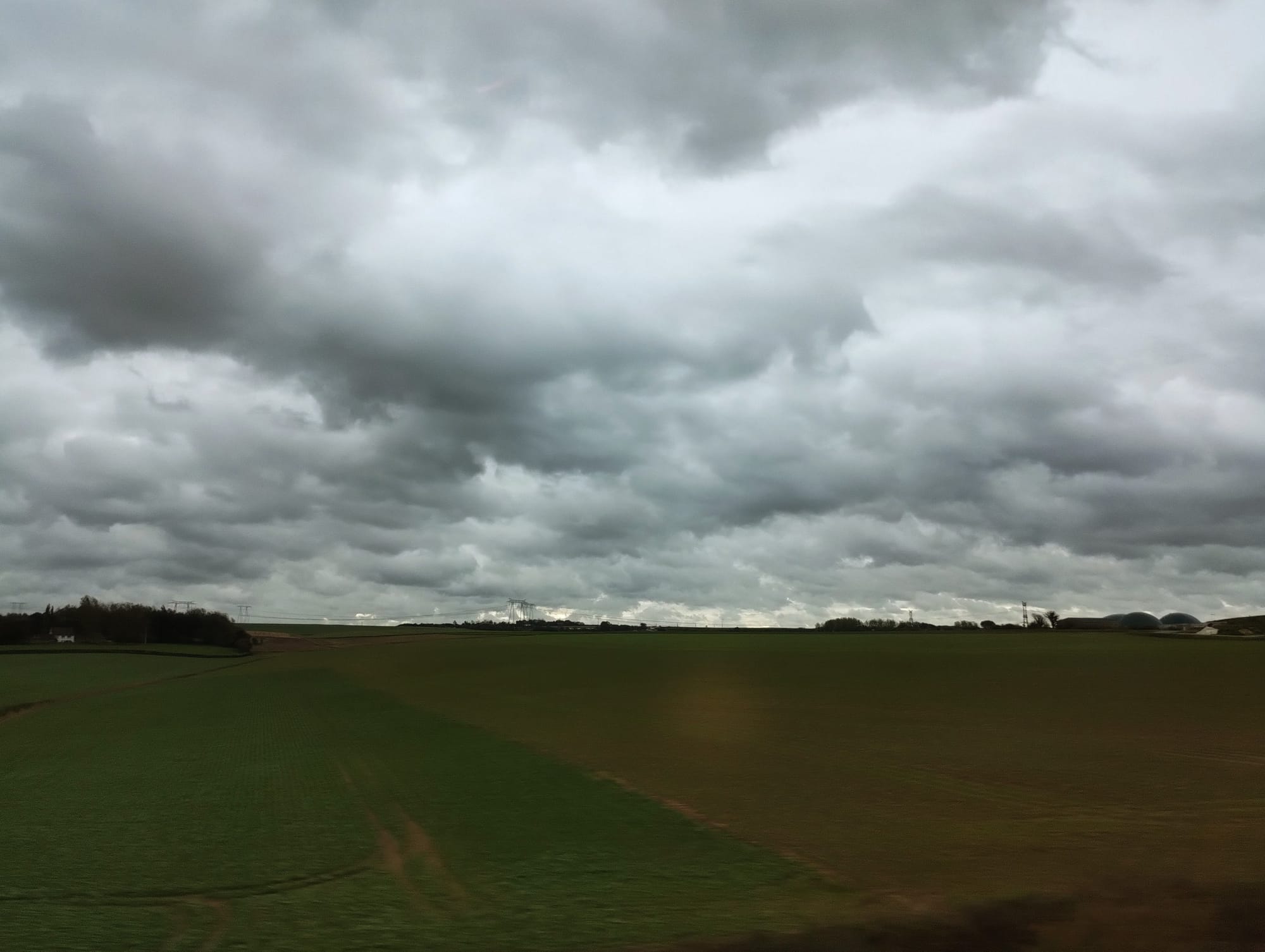 Overcast sky filled with dark, heavy clouds above a patchwork field of green and brown hues. In the distance, a treeline and power lines are visible.