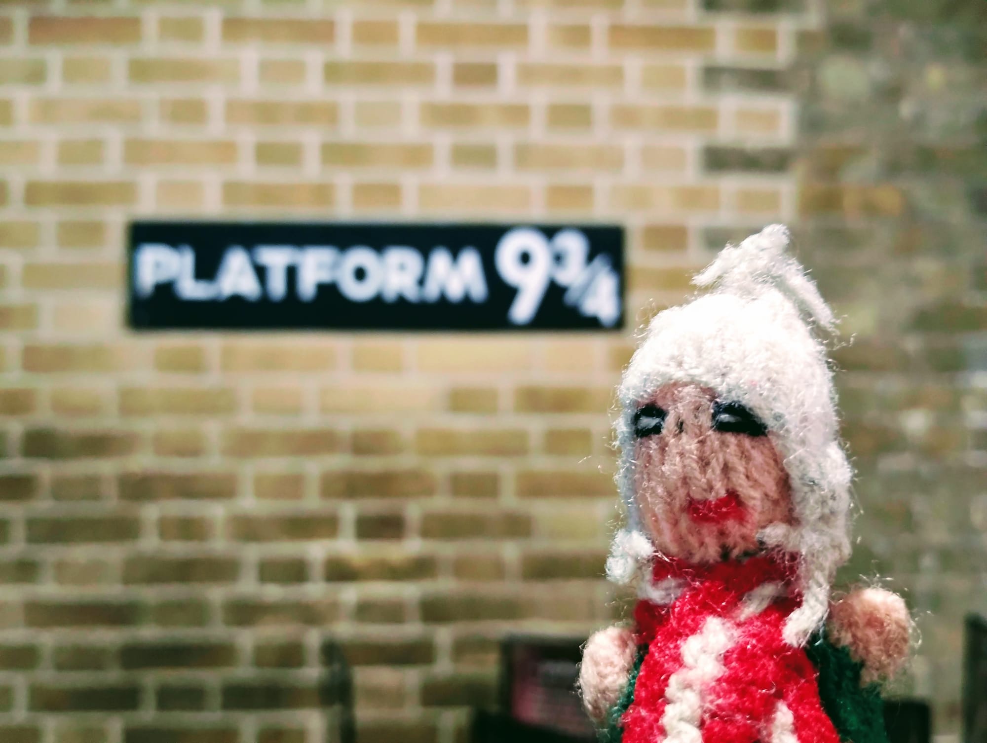 An enchanted knitted finger puppet in front of a brick wall, with a sign that reads, “PLATFORM 9 ¾”.
