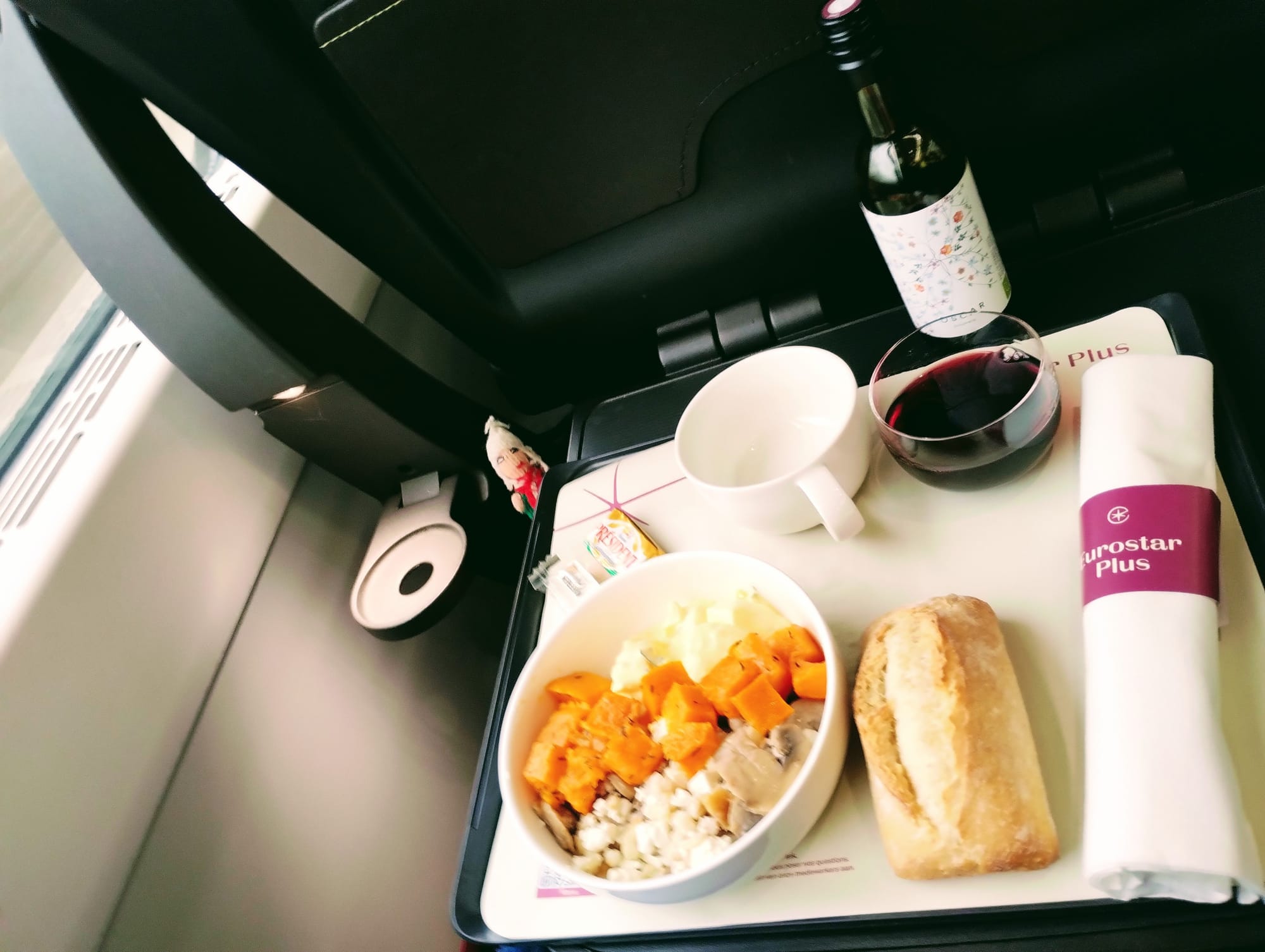 A Eurostar Plus meal tray: diced vegetables, a bread roll, red wine, tea, a mini wine bottle, and a napkin. A finger puppet admires the feast.