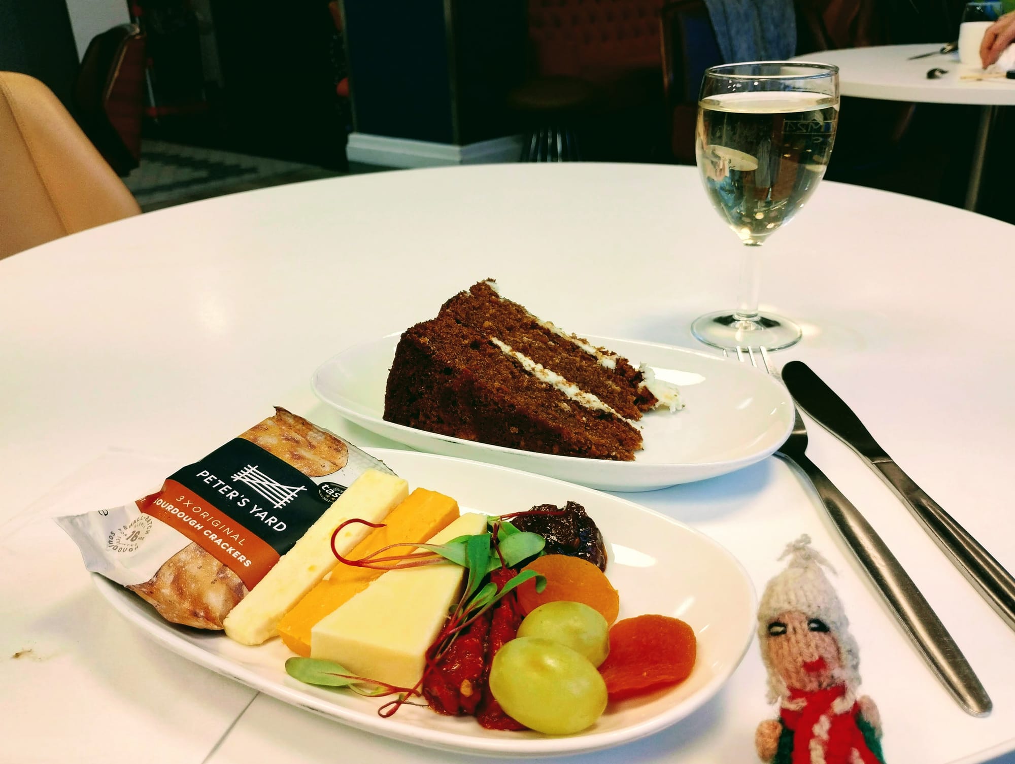 A white table setting with a slice of carrot cake, a second plate with crackers, cheese, grapes, dried apricots, and green leaves, a generous glass of white wine, and a small gluttonous finger puppet.
