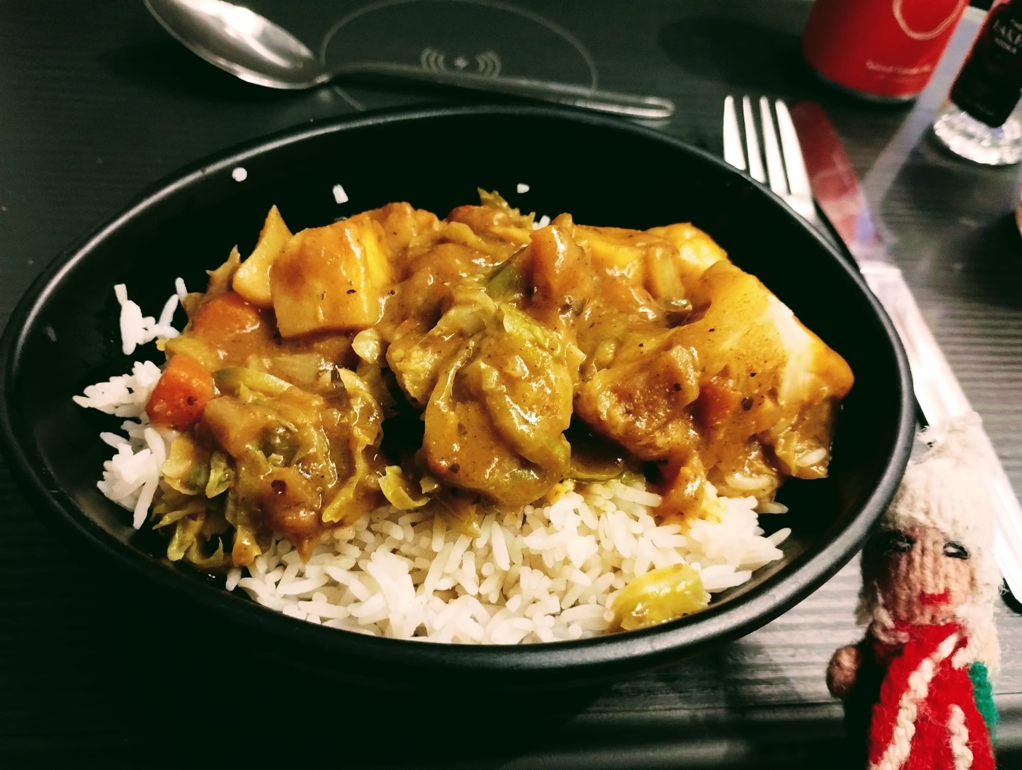 A black bowl filled with white rice, topped with a yellow-brown vegetable curry, next to a silver spoon, fork, knife, and a small finger puppet.