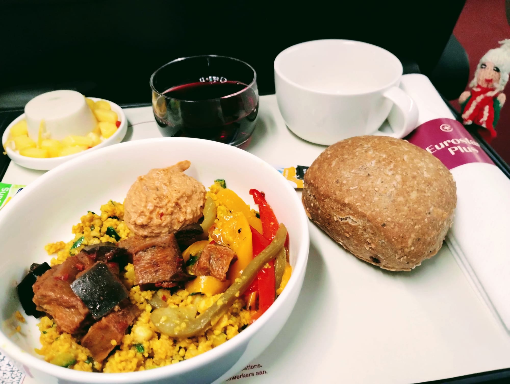 A puppet next to a meal tray with a small white bowl of couscous with grilled vegetables and curried aubergine, a bread roll, a glass of red wine, and a small dessert with fruit. A Eurostar Plus napkin holds cutlery.