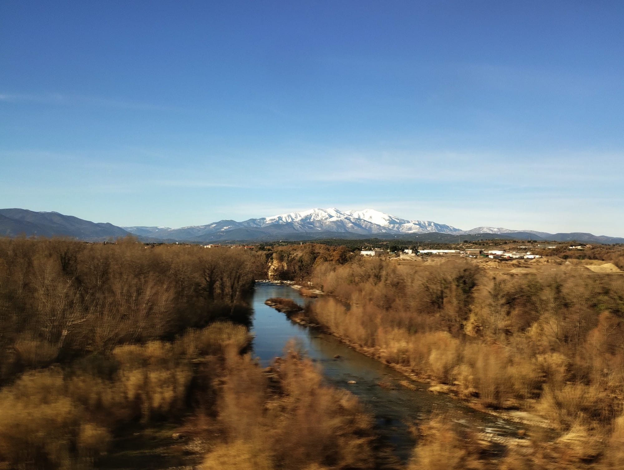 River flowing through a valley, flanked by bare trees and leading to snow-capped mountains under a clear blue sky.