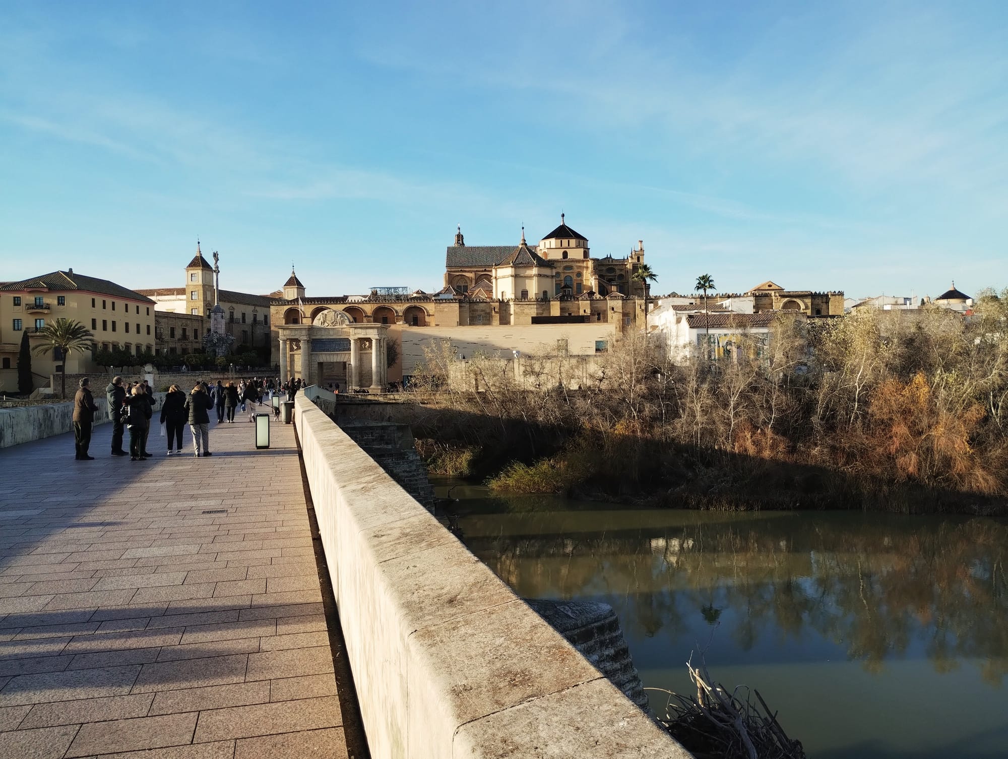 View from the stone bridge across the river towards the Mezquita-Cathedral of Córdoba, a large beige and brown architectural complex with multiple domes and structures, under a clear blue sky. People walk along the bridge towards the cathedral, with low wintry trees lining the riverbanks.