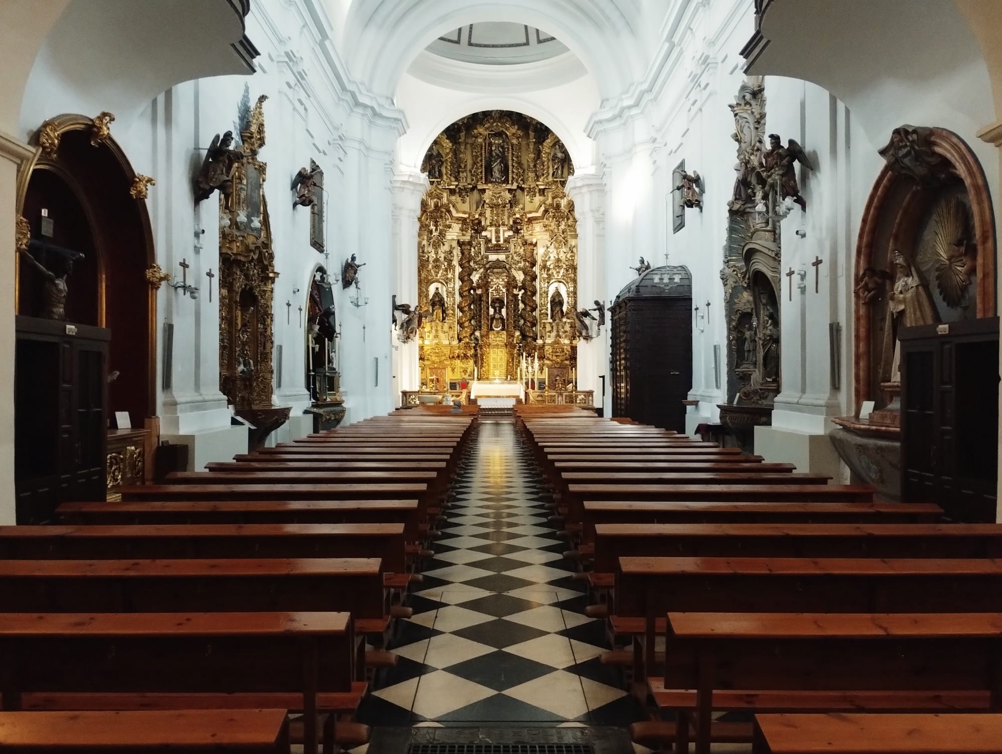 Interior view of the San Juan y Todos Los Santos church, centred on rows of wooden pews facing a gilded altar at the far end, with a check black and white floor pattern leading the eye. The white walls are adorned with sculptures and statues.
