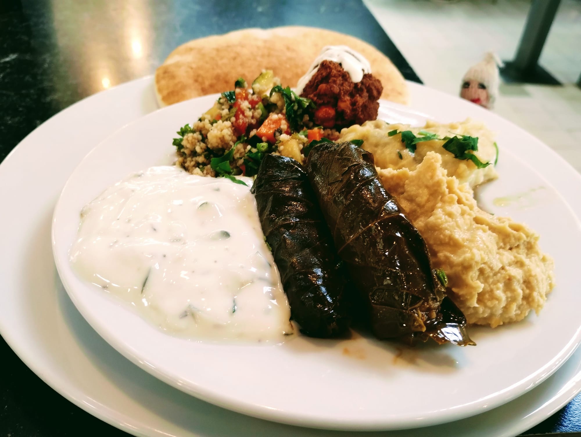 A plate of assorted Middle Eastern food including stuffed grape leaves, hummus, baba ghanoush, tabouleh, and a falafel ball with pitta bread.