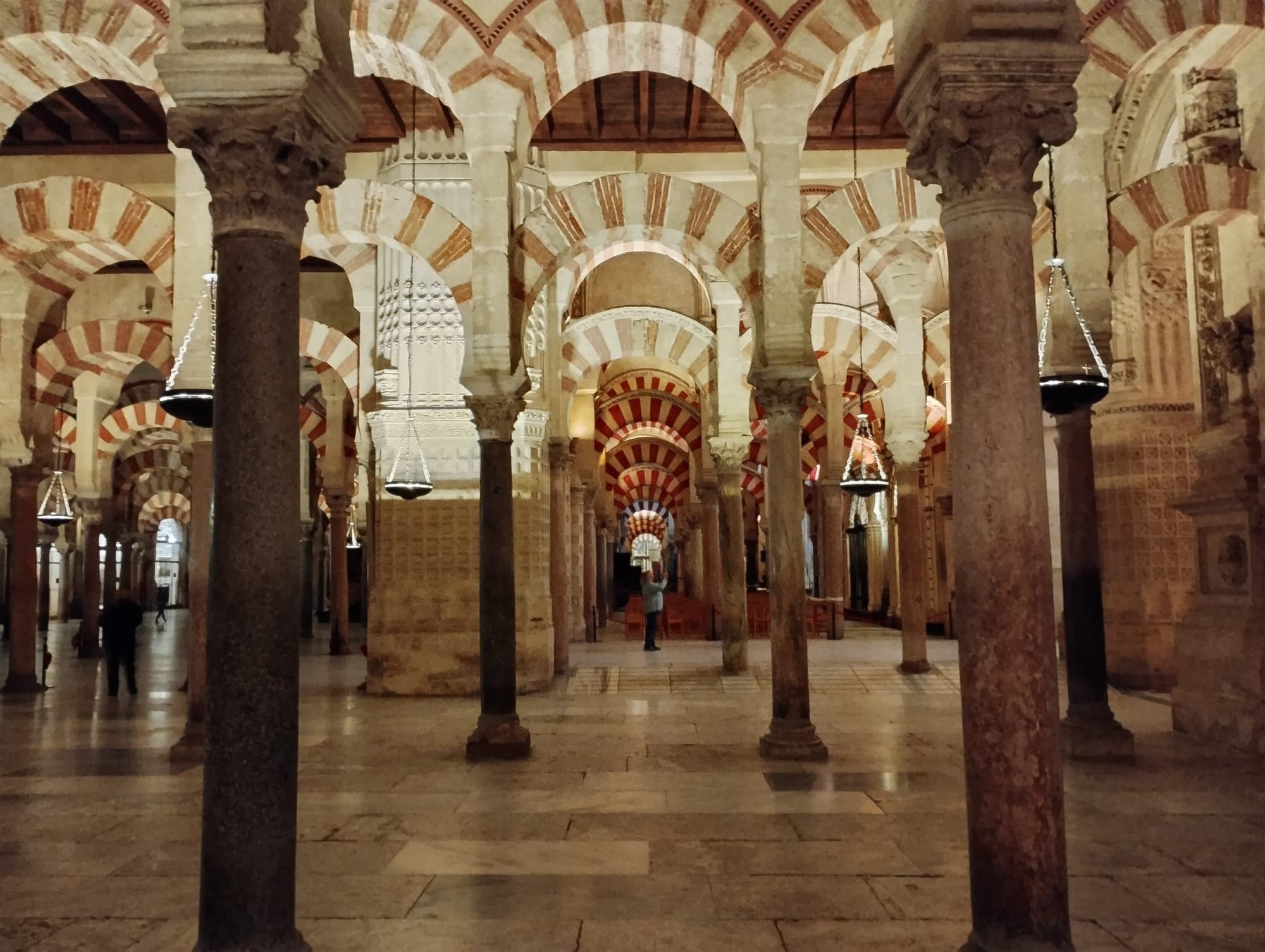 Interior view of the Great Mosque of Cordoba, Spain, featuring rows of striped red and white arches supported by dark stone columns, ornate detailing, and hanging lamps, leading the eye into the distance where a person is standing, taking a photograph.