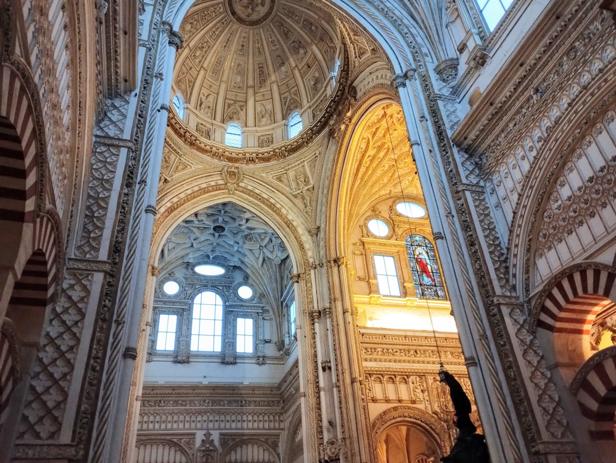 Interior of the Mezquita-Cathedral of Cordoba, Spain, showcasing ornate, arched ceilings and walls with detailed carvings, columns, and windows, the focus on the upper levels of the cathedral. A large dome is visible in the background, a stained-glass window with a red figure is on the right, and the light from the windows creates a contrast of light and shadows within the space.