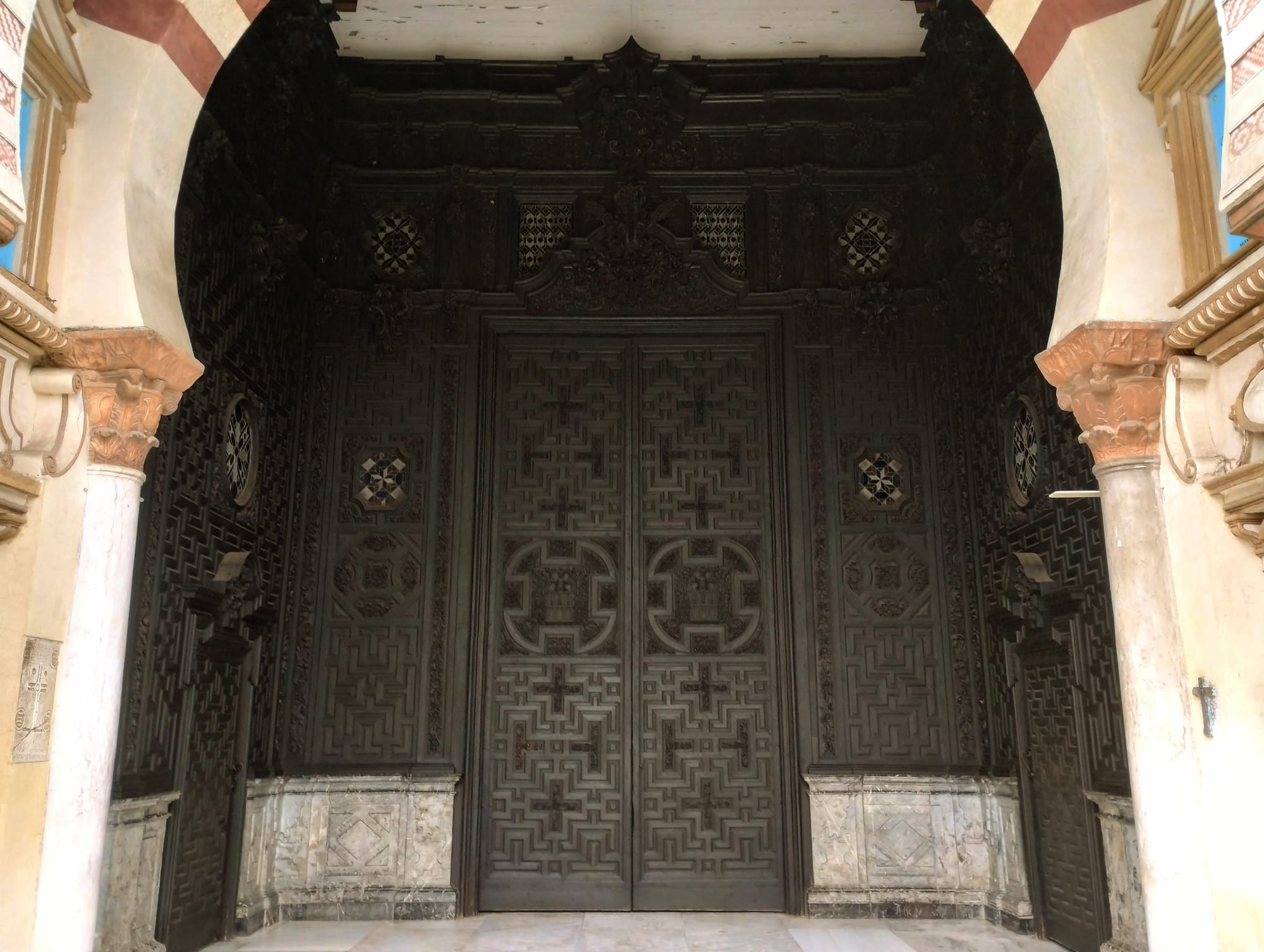 The Puerta de Las Palmas, dark wooden double doors with intricate geometric carvings and decorative panels, recessed within a grand arched entrance, supported by ornate columns in a horse-shoe arch.