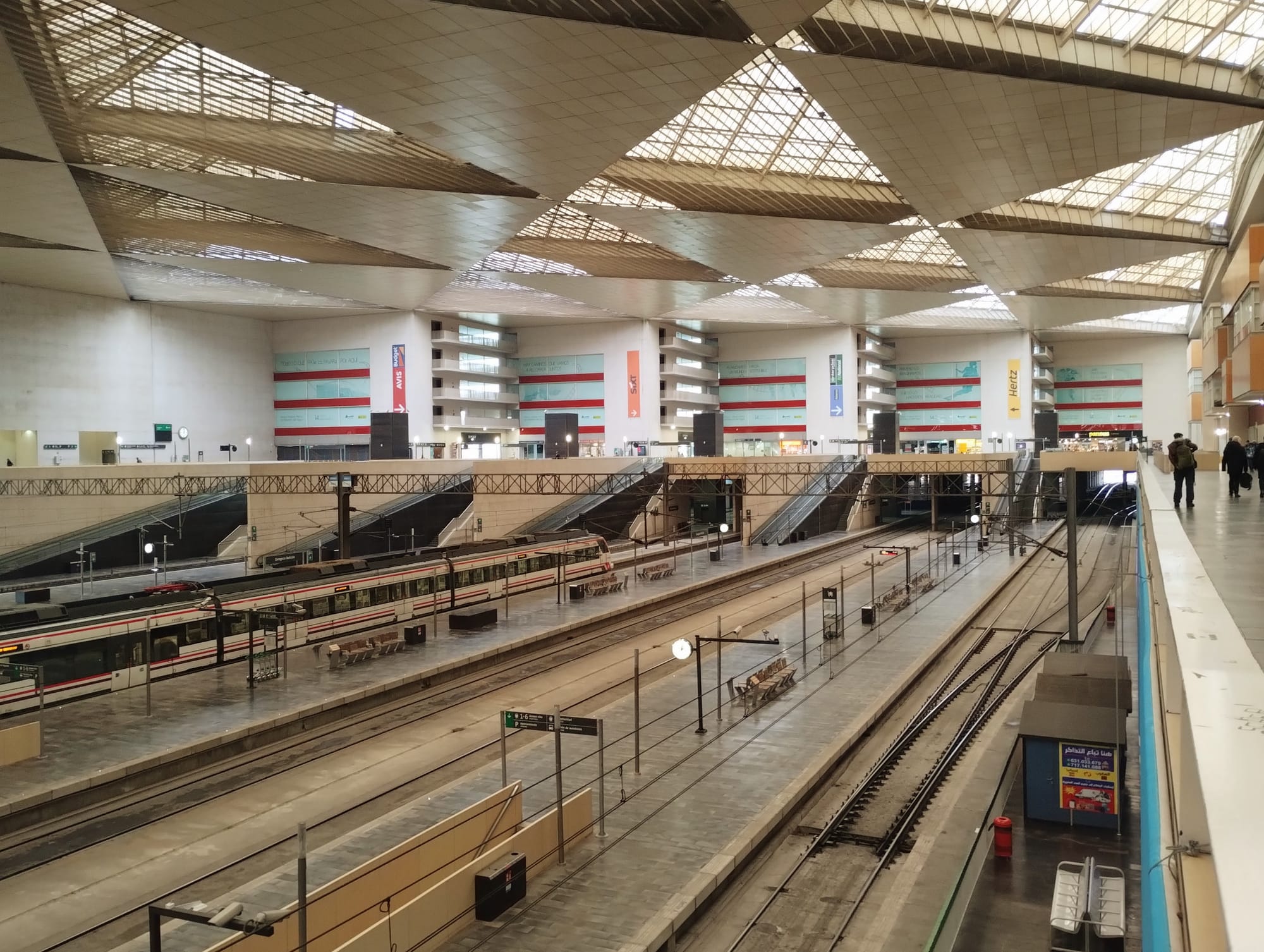 Interior view Zaragoza Delicias railway station with multiple platforms and tracks, featuring a geometric, translucent ceiling. A train is at a platform, and escalators lead up to upper levels. Signage for car rental and services is visible on the walls. People are walking on the upper level.