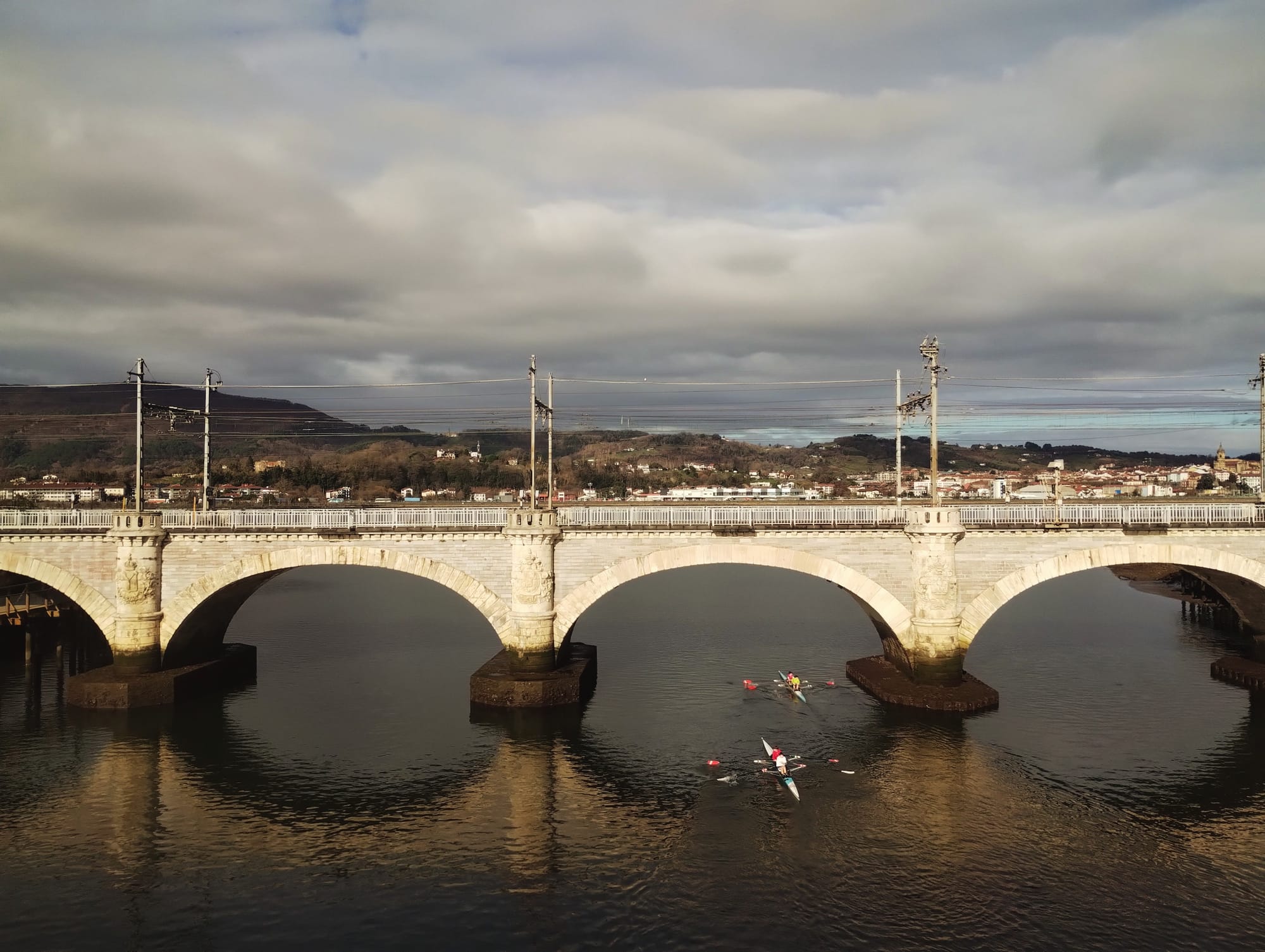 A stone arch bridge spanning a river, with two rowing teams in the water below. The bridge is composed of three large arches, with elaborate details on the piers and a railway line above. The river water reflects the bridge and cloudy sky. The backdrop features a distant town nestled on a hillside under a cloudy sky.