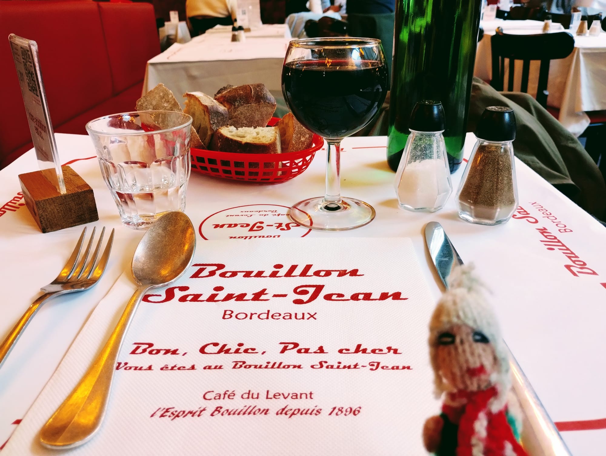 Finger puppet waiting at a table setting in a French restaurant, featuring a wine glass filled with red wine, a bread basket, glassware, salt and pepper shakers, silverware, and a napkin with the restaurant's name "Bouillon Saint-Jean" in red.