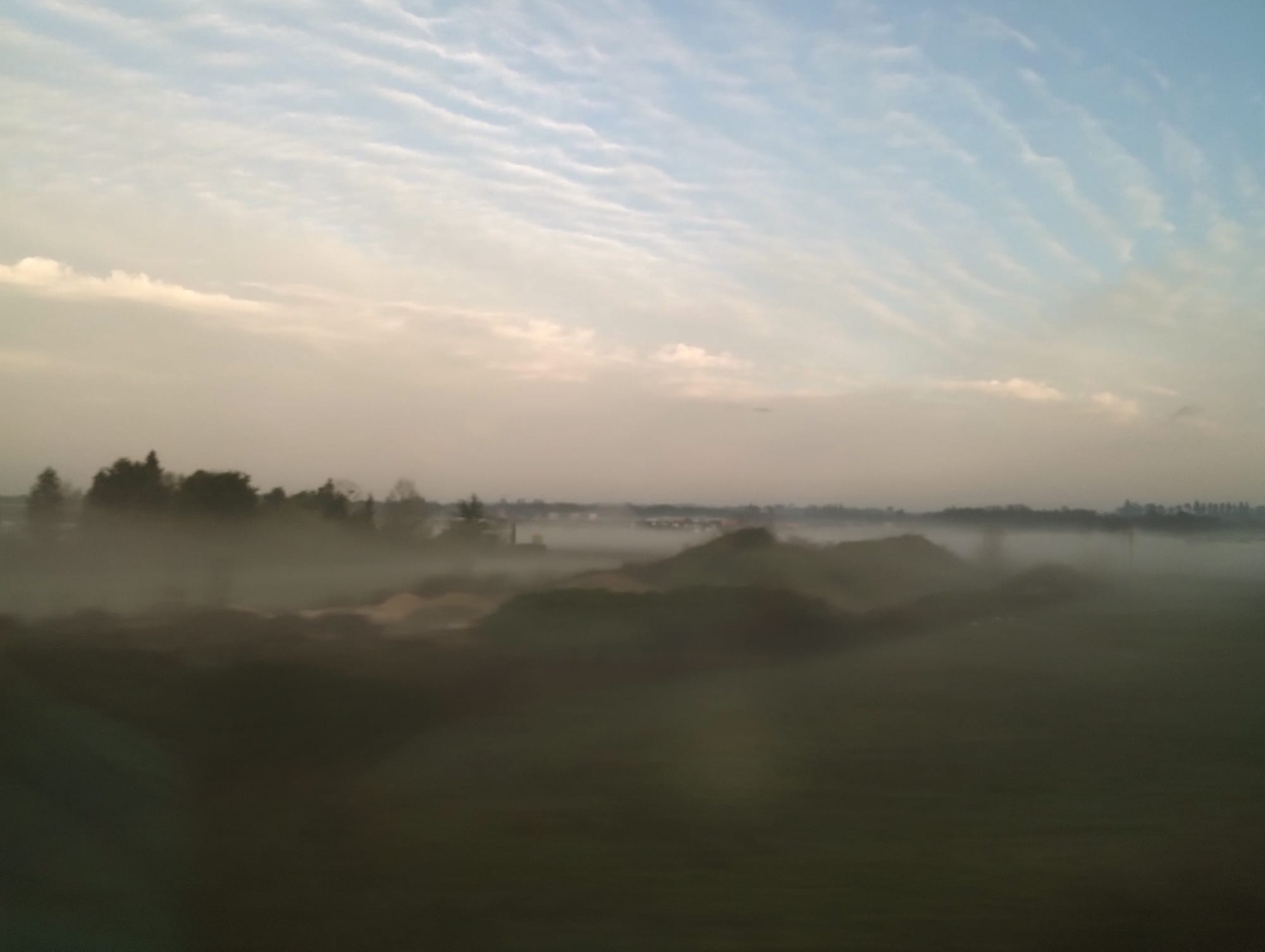 Landscape with low-lying fog covering a rolling field and treeline, a light blue sky with wispy clouds visible above.