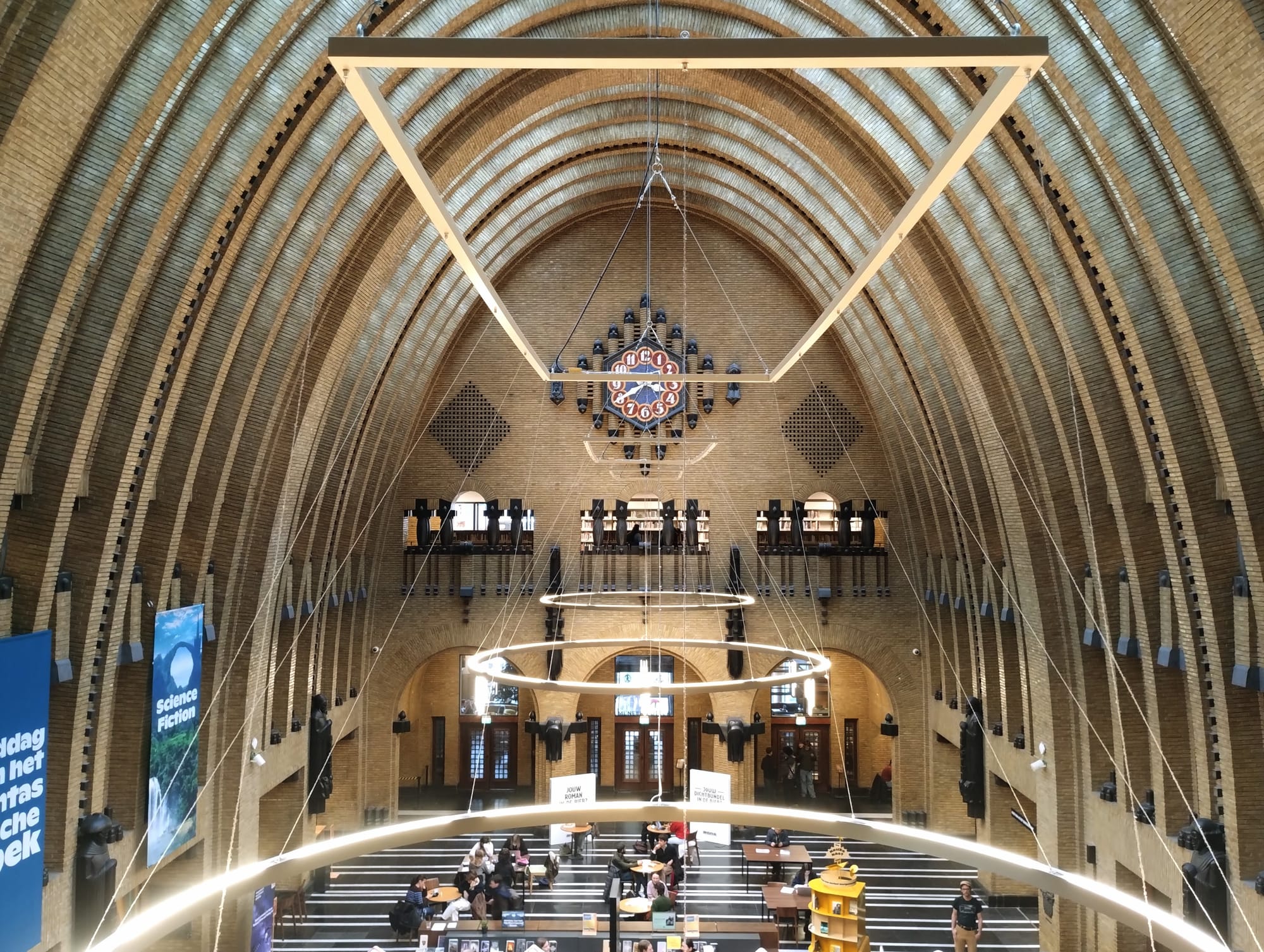 A wide-angle shot of a library with an arched brick ceiling, geometric light fixtures, and multi-level seating area. A decorative clock above a central entrance is on the far wall, with a banner for 'Science Fiction' on the left.