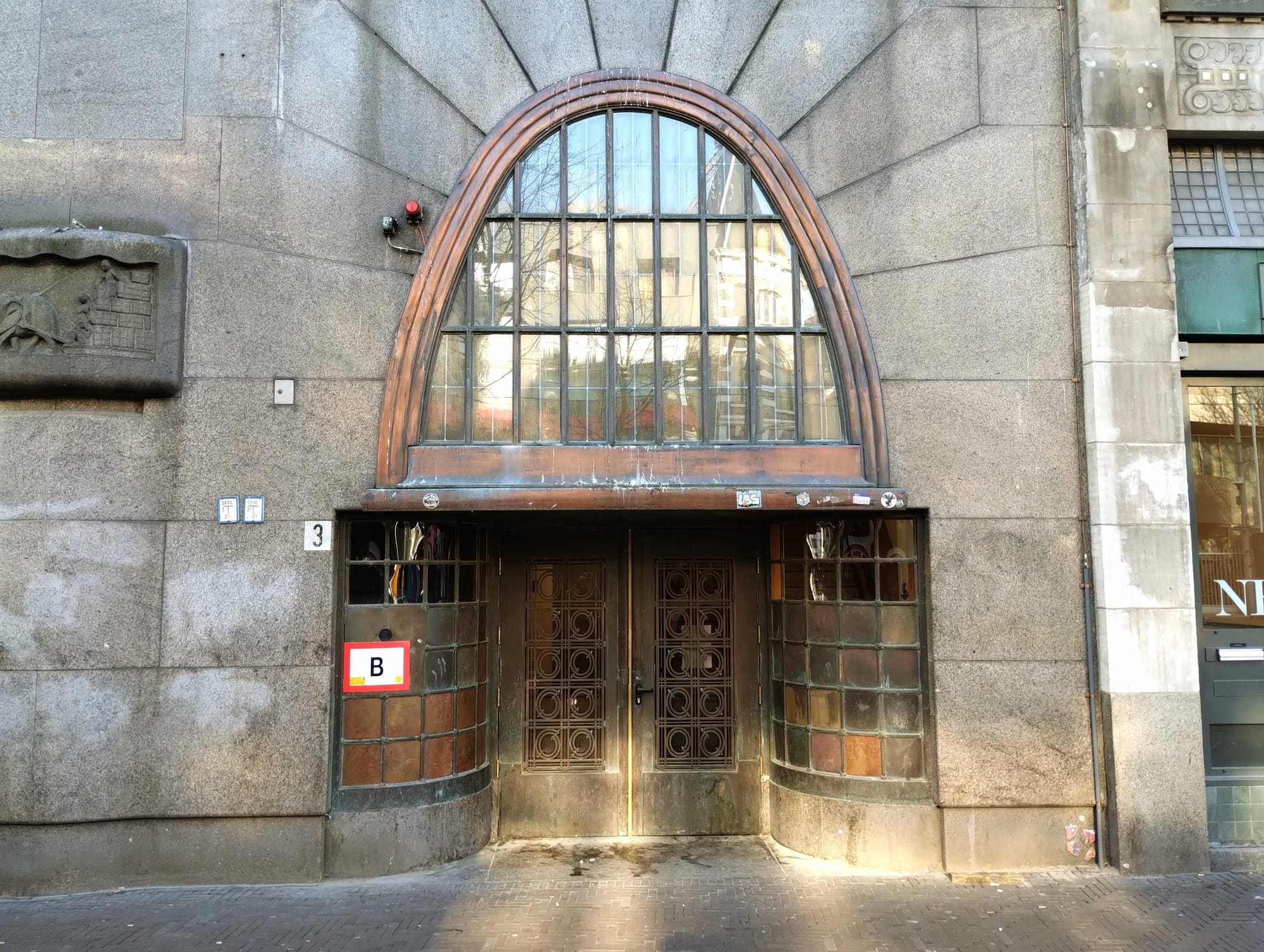 An arched doorway with a large, multi-paned window above two bronze double doors, set into a stone building facade, with a side window and a "B" sign next to the doors.