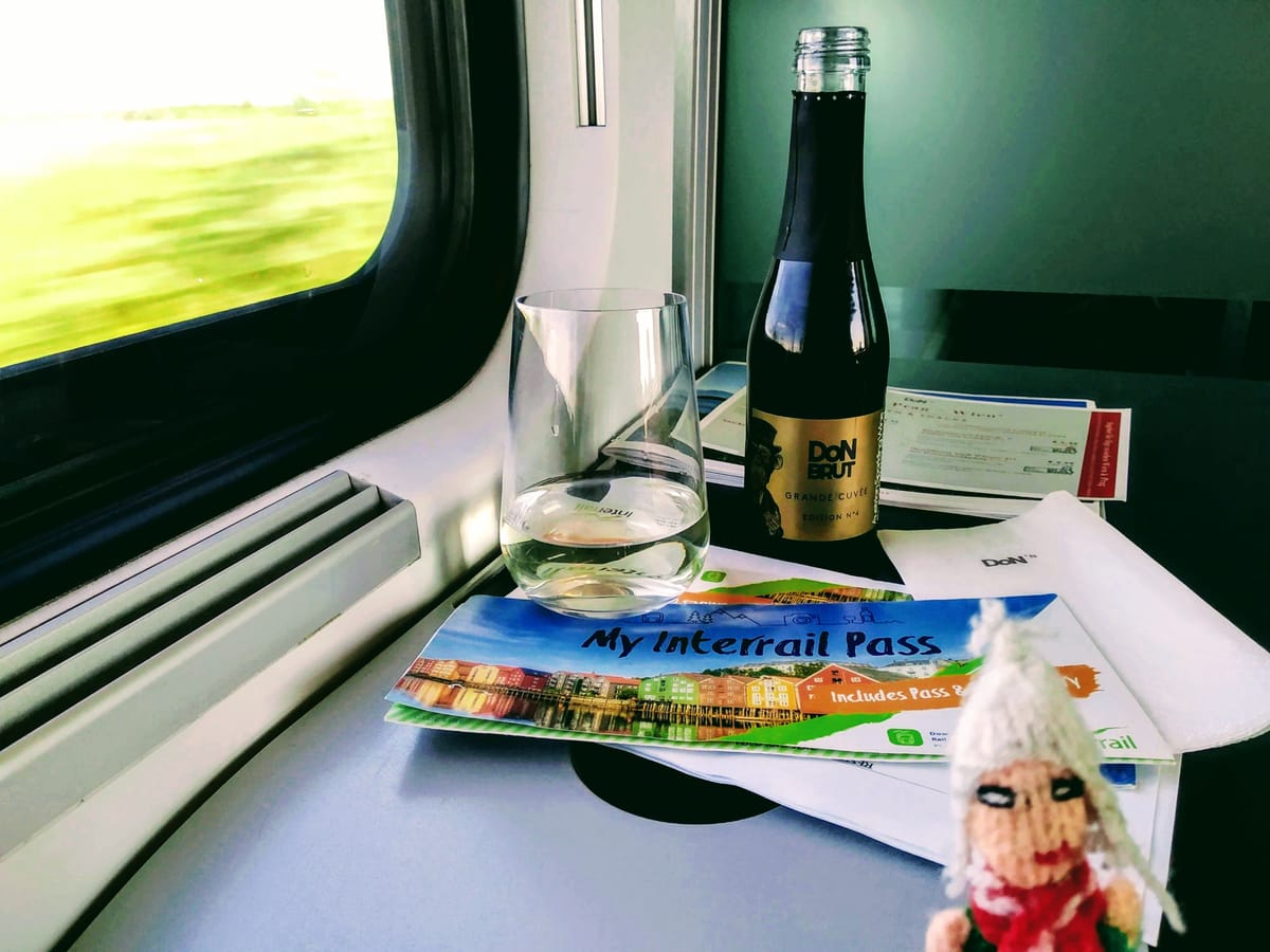 A fearless Peruvian finger puppet inside a moving train examines a small bottle of sparkling wine and an Interrail pass, while blurred green landscape passes the window.