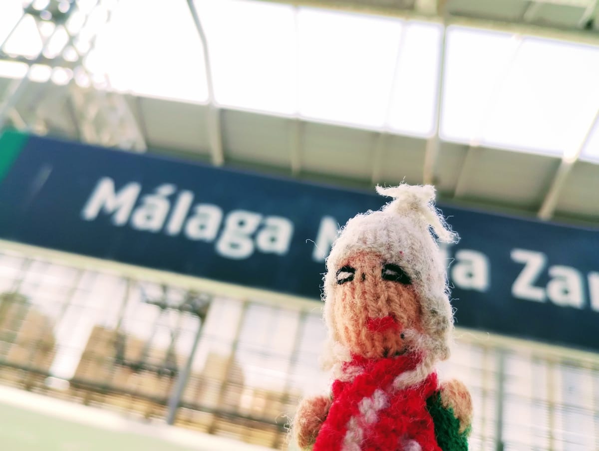 A jaunty Peruvian finger puppet stands in front of the blue platform sign at Málaga María Zambrano train station.