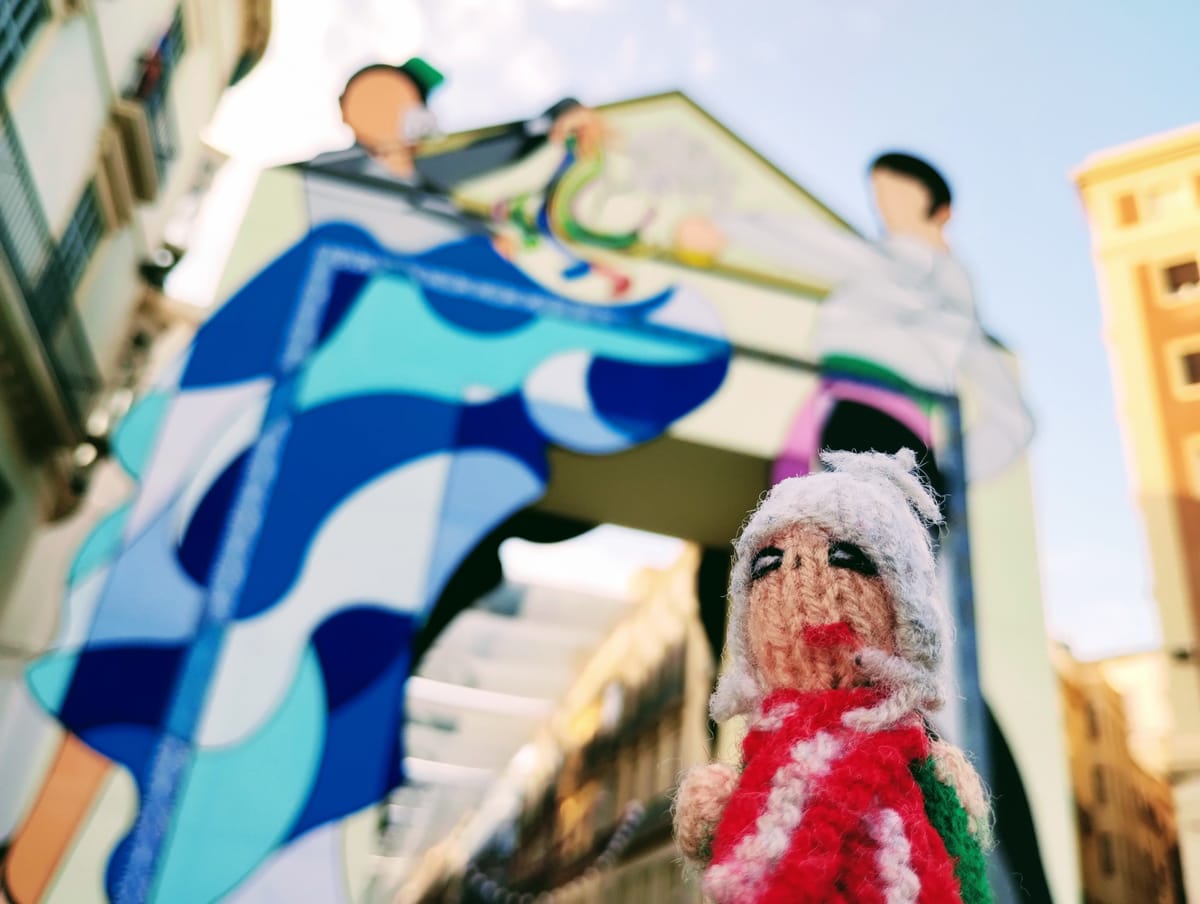 A feisty finger puppet with red and white dress bristles in front of a building painted with a mural of abstract tango figures.