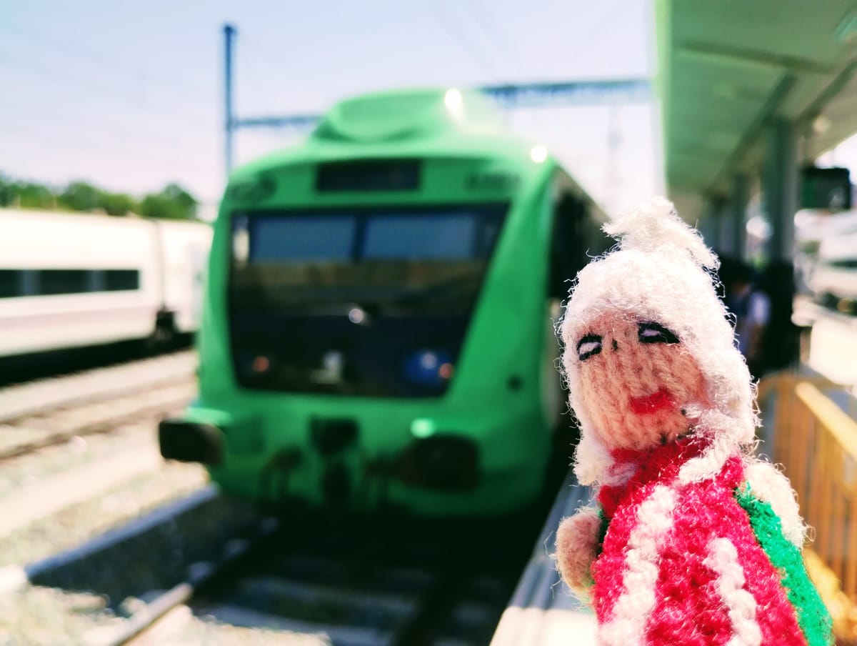 A coquettish finger puppet standing on a train platform with a bright green train in the background.