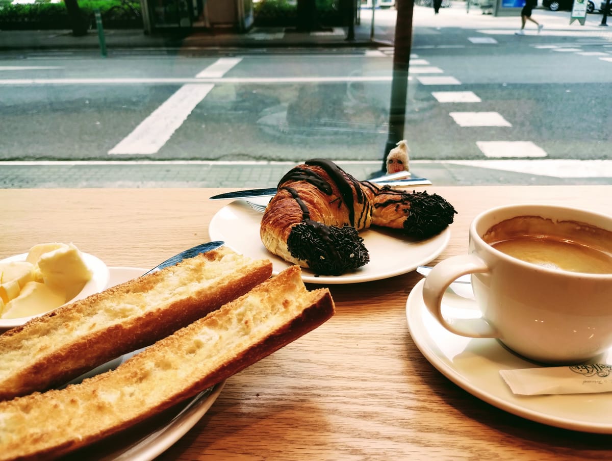 From behind a wooden table and in front of a window looking onto a Donostian street, a finger puppet surveys a breakfast of toast and butter, a chocolate croissant, and a cup of coffee.