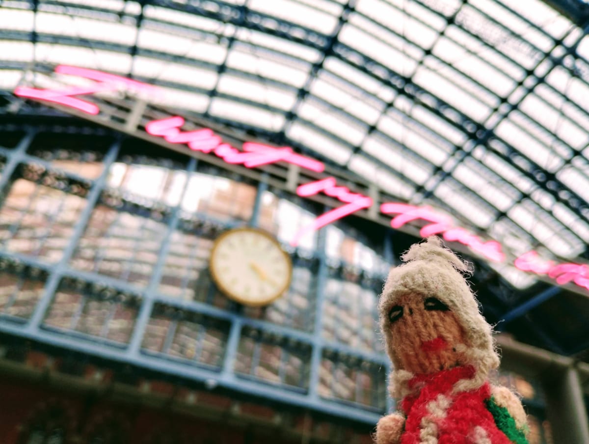 A Peruvian finger puppet bristles under the clock in London Saint Pancras International. Above him is written "I want my time with you" in neon pink.