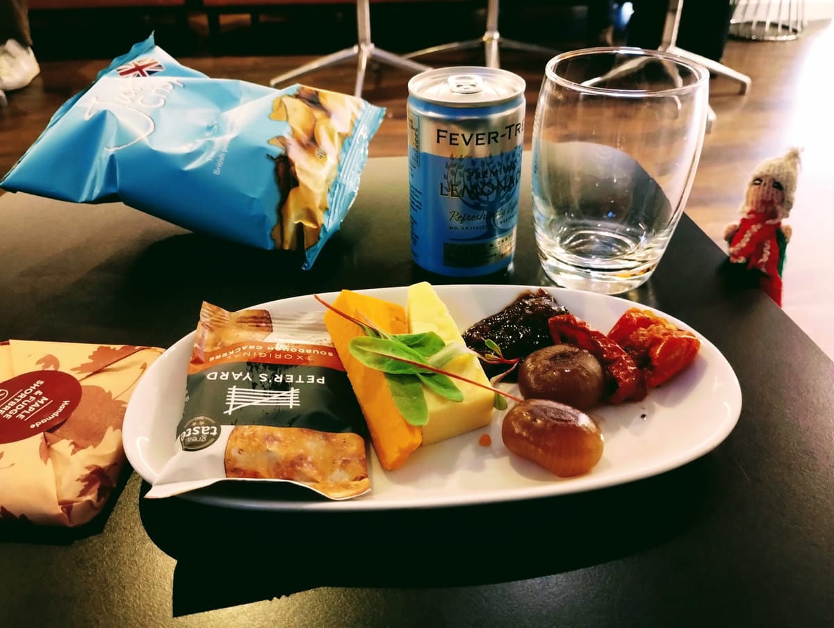 A self-indulgent finger puppet surveys his in-lounge spoils: cheese, pickled onions, crackers, shortbread, a can of Fever-Tree, and a bag of crisps.