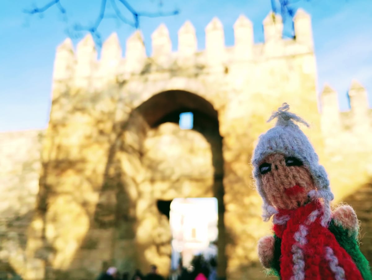 A Moorish-feeling finger puppet in front of a large, weathered stone archway into Córdoba's old town, under a blue sky.