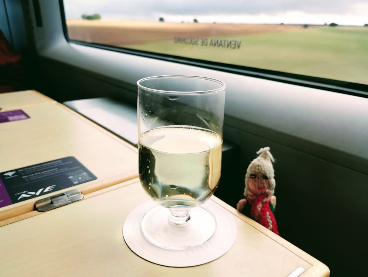 Finger puppet proudly showing off a glass of sparkling white on a white coaster on a train table. A blurred view of fields and a cloudy sky through the window.