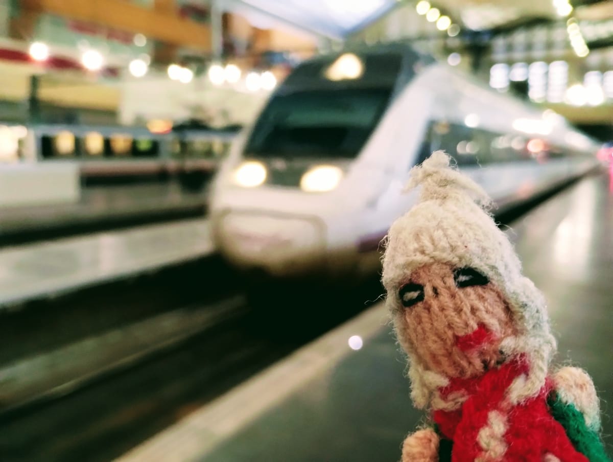 Rugged Finger Puppet wearing a hat and scarf, ready to board the approaching train on a platform in Zaragoza Delicia station.