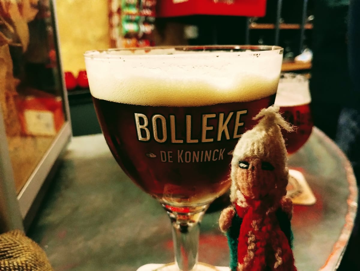 An erect Peruvian finger puppet stands in front of a tulip-shaped glass filled with dark amber beer, topped with thick, foamy head. "Bolleke De Koninck" is written on the glass.