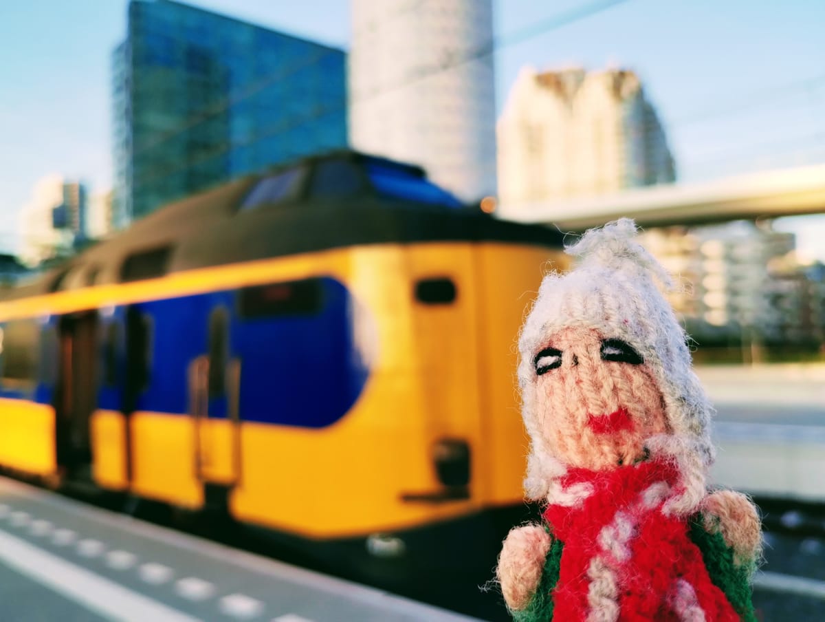 Finger Puppet stands on a platform in front of a yellow and blue NS train, with Den Haag blurred under a blue sky in the background.